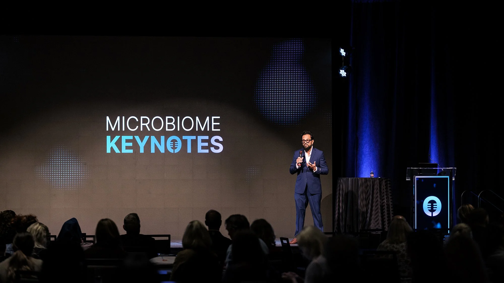 A man in a blue suit speaking into a microphone on stage at a conference titled 'Microbiome Keynotes,' with an audience in front and large screen behind him displaying the same text.