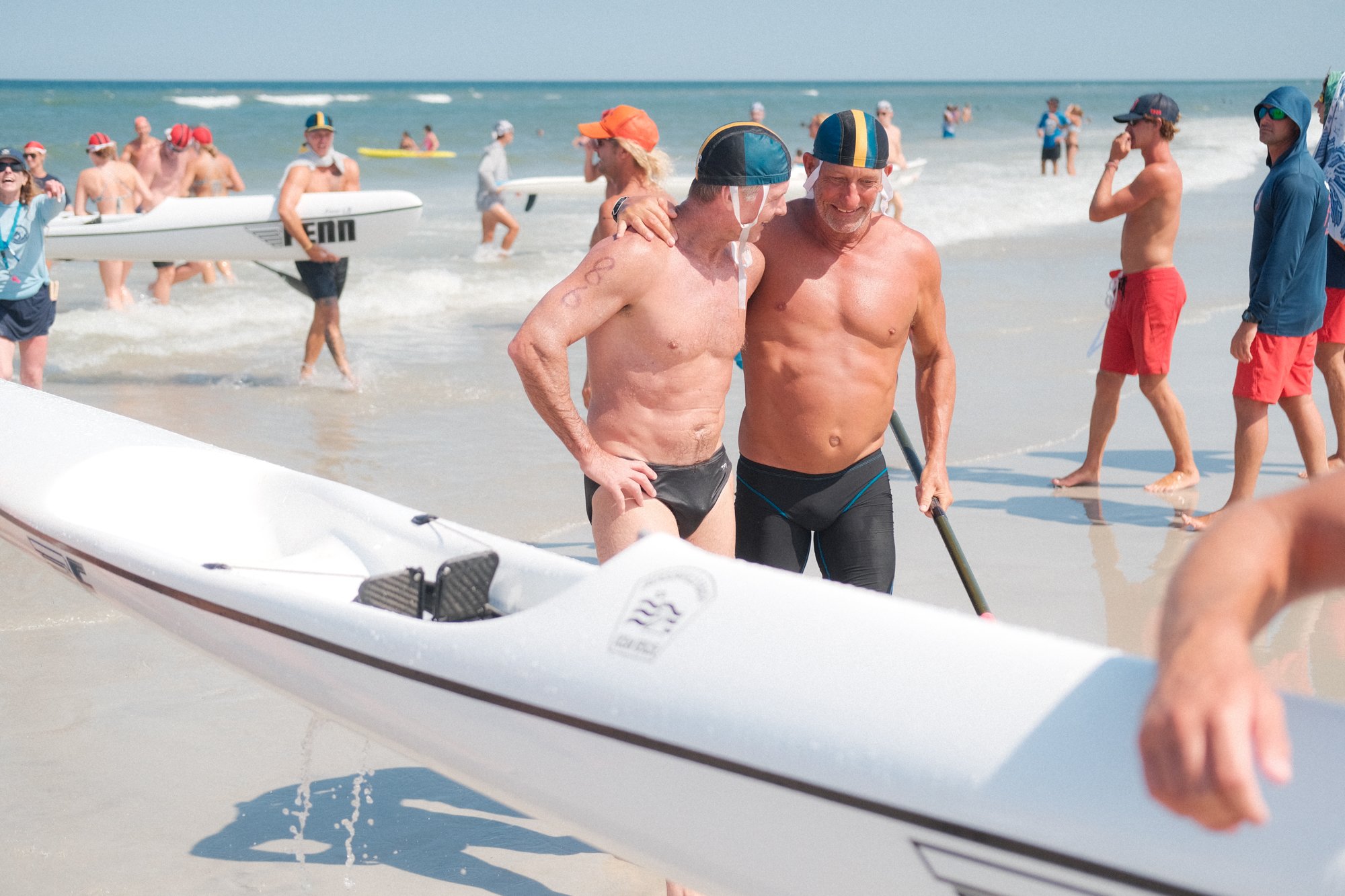 Two shirtless older men with swim caps and wetsuits around their waist smiling and talking on a beach with many people in the water and some holding surfboards in the background.