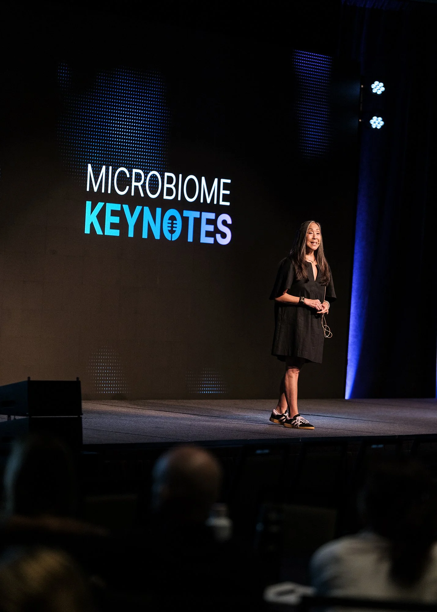 A woman standing on a stage, speaking at the Microbiome Keynotes event, with a large screen behind her displaying the event name and logo.