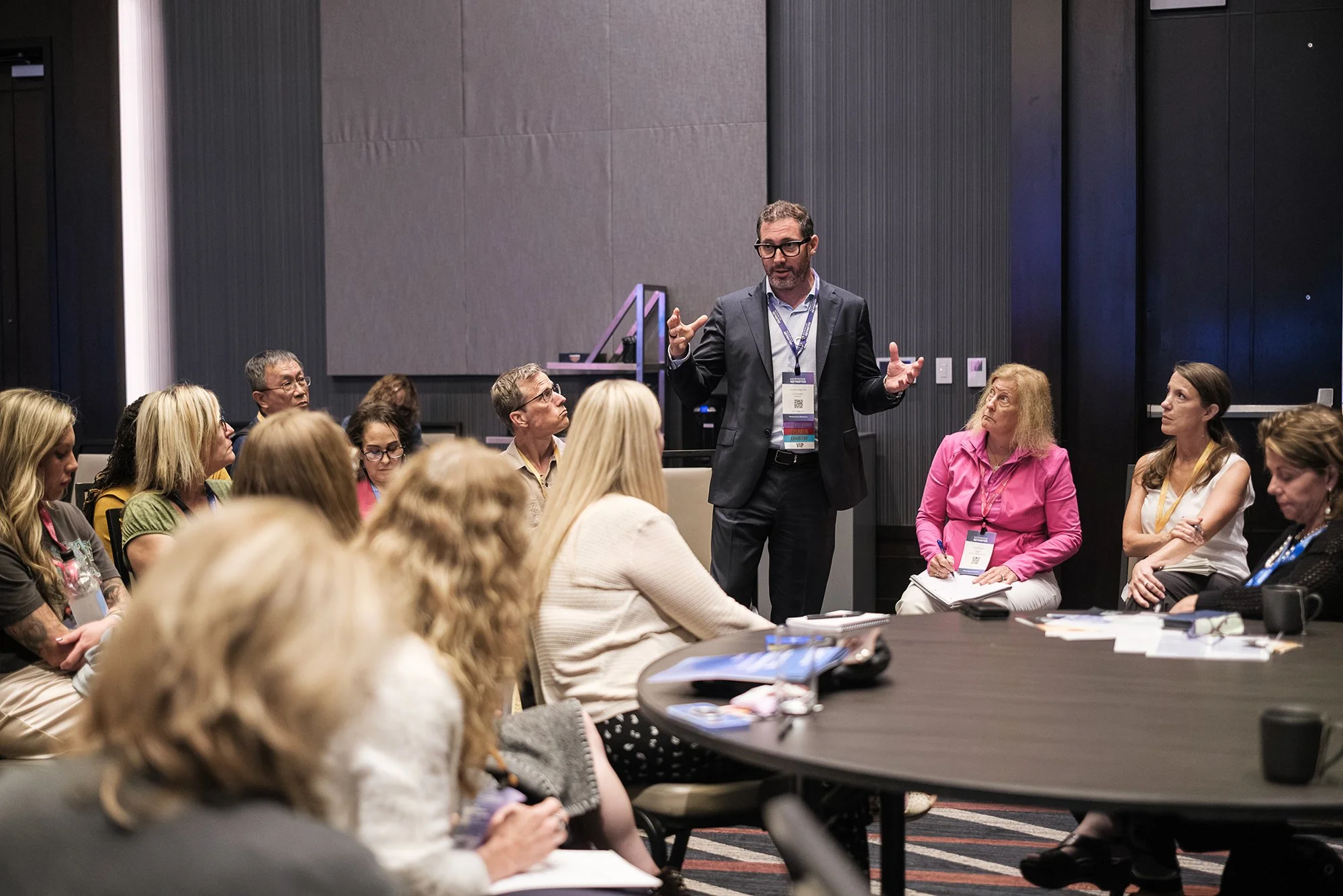A man in a suit stands and speaks to a group of seated people at a conference. The audience listens attentively, some taking notes, in a modern indoor setting.