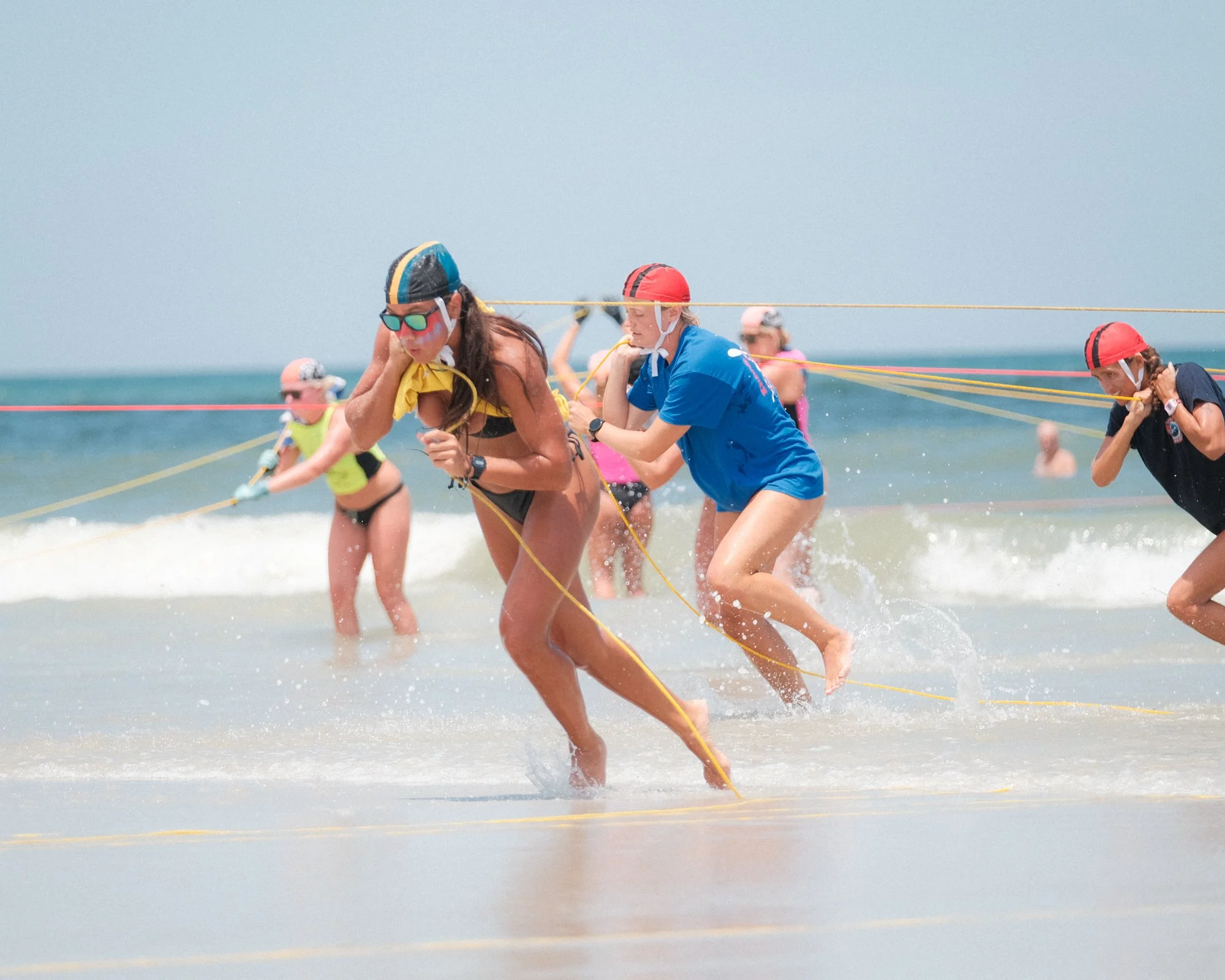 People participating in an outdoor race at the beach, running through shallow water with yellow racing ropes and wearing hats and sunglasses.