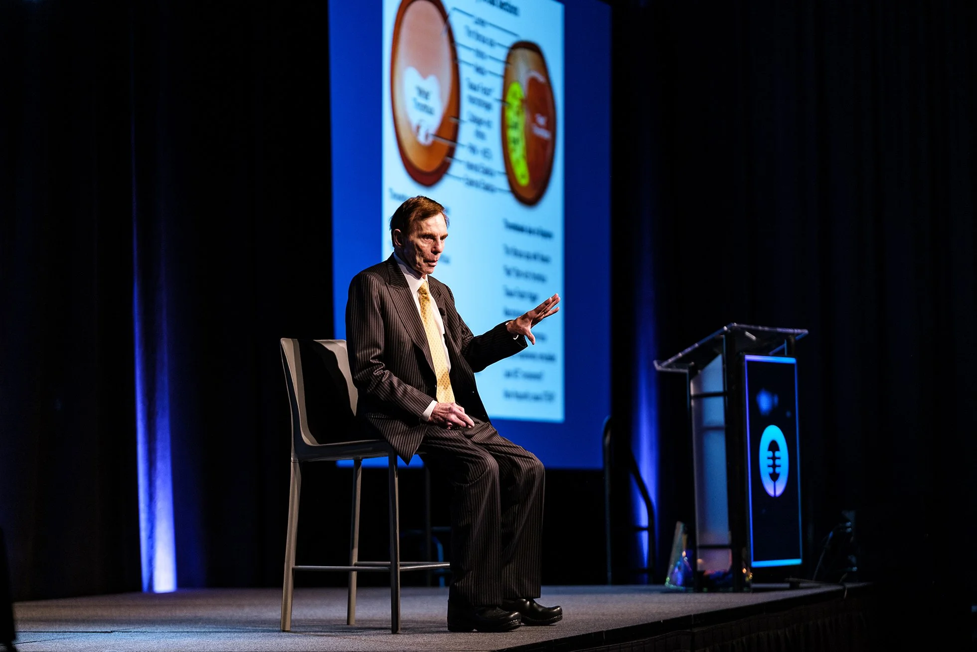 A man in a pinstripe suit sitting on a chair on a stage, gesturing with his right hand while speaking. Behind him is a large screen displaying a diagram or presentation slide with a circular chart. The scene is lit with blue accent lighting.