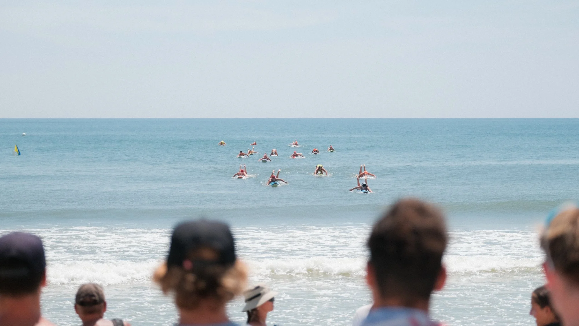 People participating in a surf competition or surfing lesson in the ocean, with spectators on the beach watching.