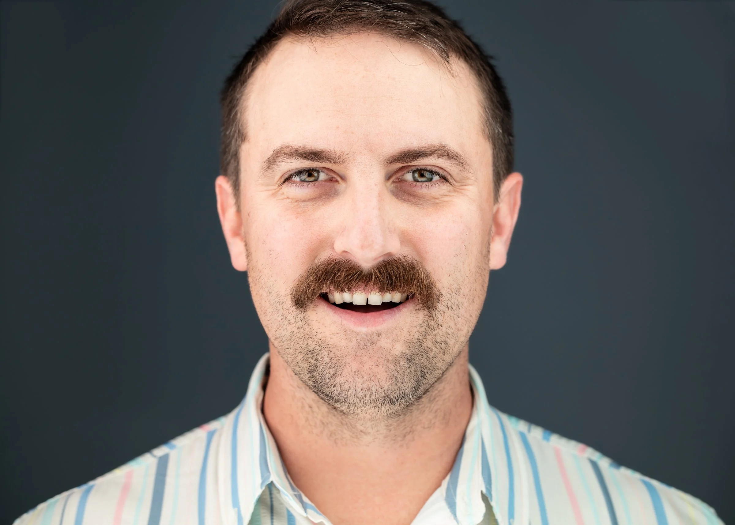 Close-up of a smiling man with a beard and mustache, wearing a striped shirt, against a dark background.