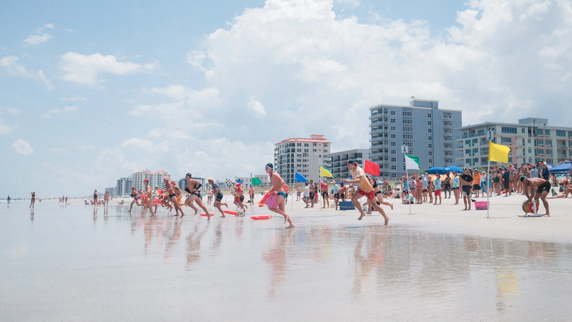 Beach scene with people swimming and running at the water's edge, with buildings and umbrellas in the background under a partly cloudy sky.