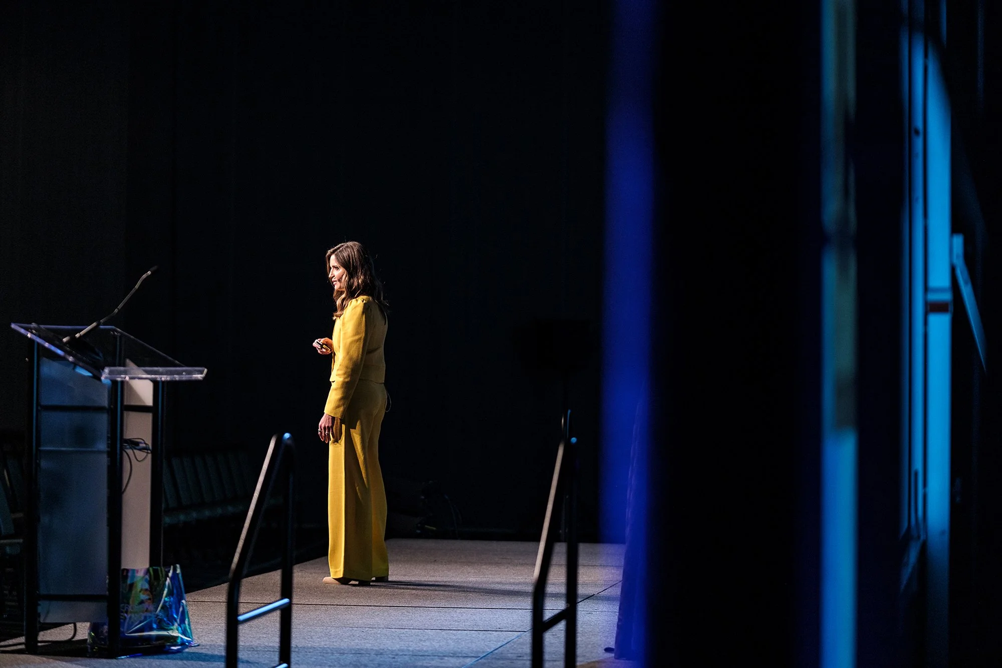 A woman in a yellow pantsuit standing on stage, smiling, holding a remote or clicker, with a podium and microphone nearby, in a dimly lit conference or presentation setting.