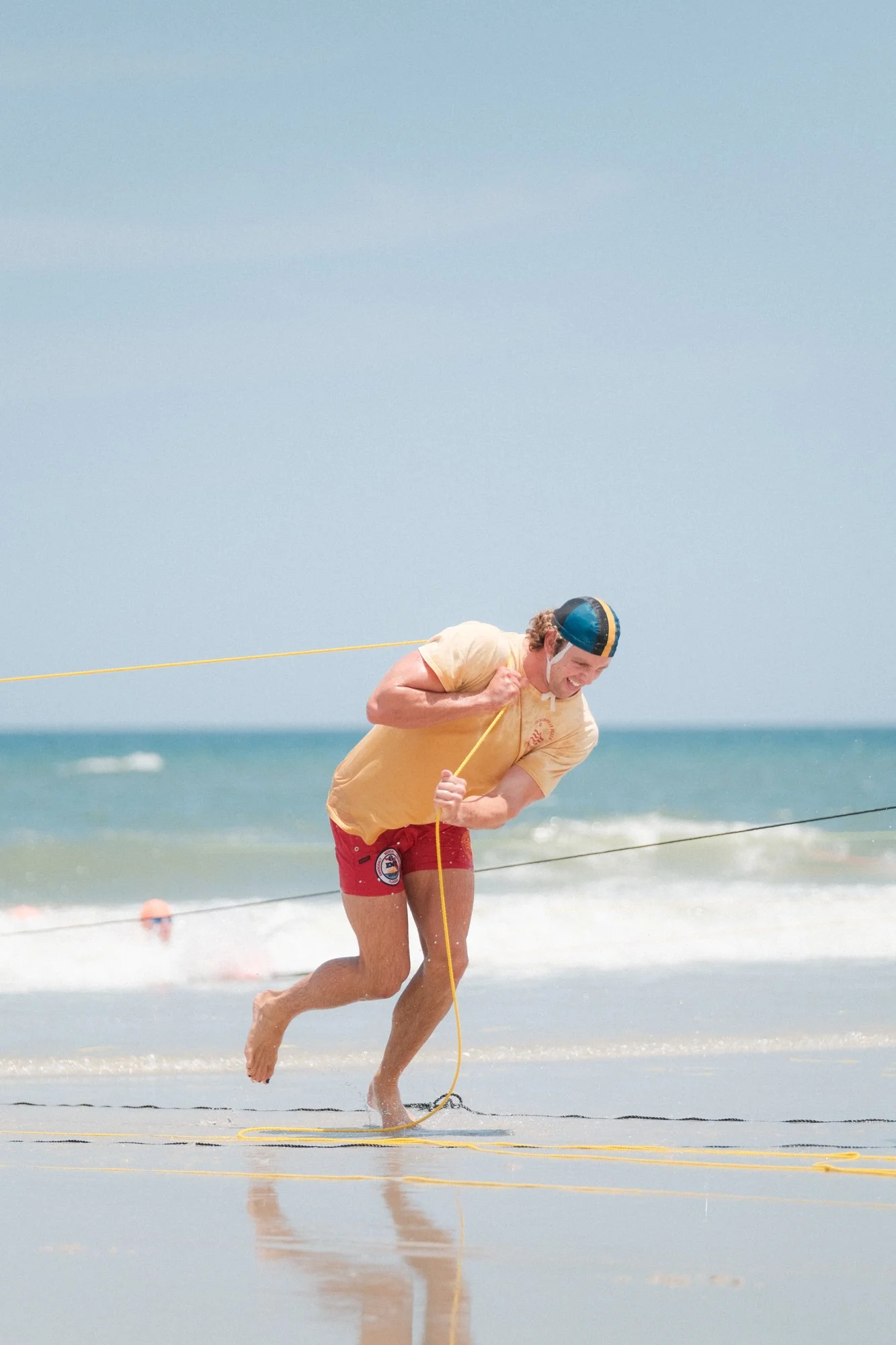 Man running on the beach holding a yellow rope, wearing a yellow t-shirt, red shorts, a cap, and a white mask, with water and a blue sky in the background.