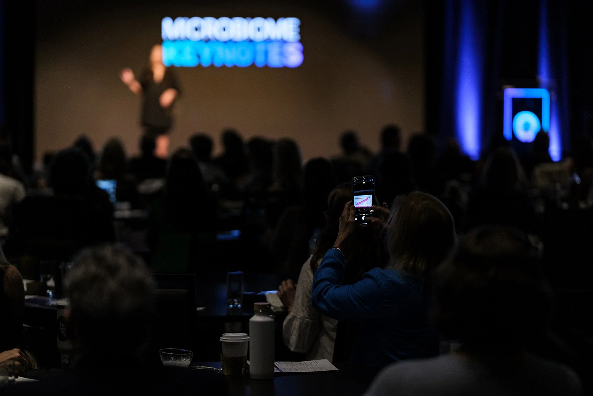 A conference room filled with seated attendees, some taking photos or looking at a speaker on stage. The stage features a blurred person standing near a large screen with bright blue text that reads "NARGARAGOME IS VOTES." The room has dark walls wit