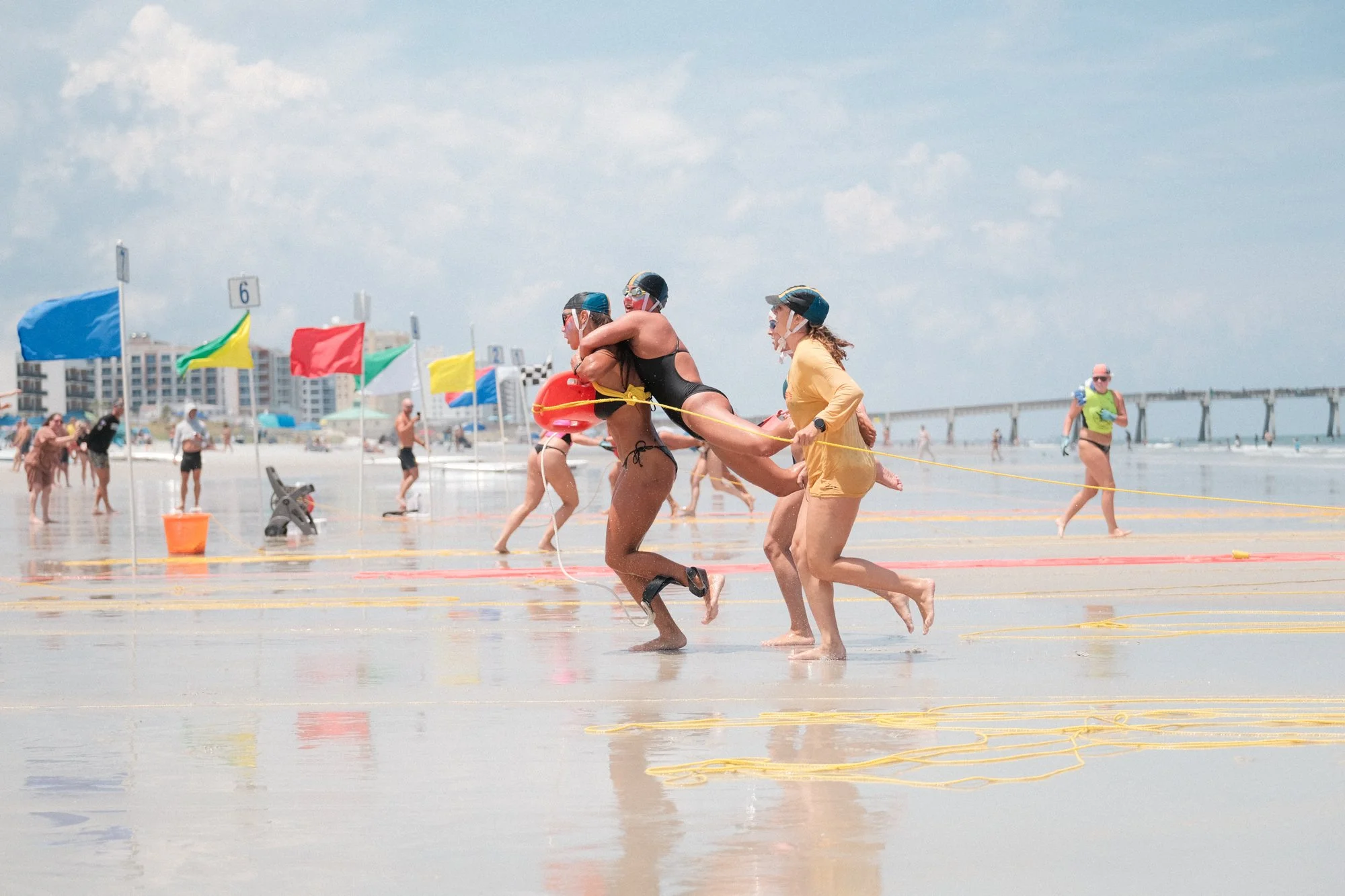People participating in a beach lifeguard race, running through shallow water with flags and beach equipment nearby on a sunny day.
