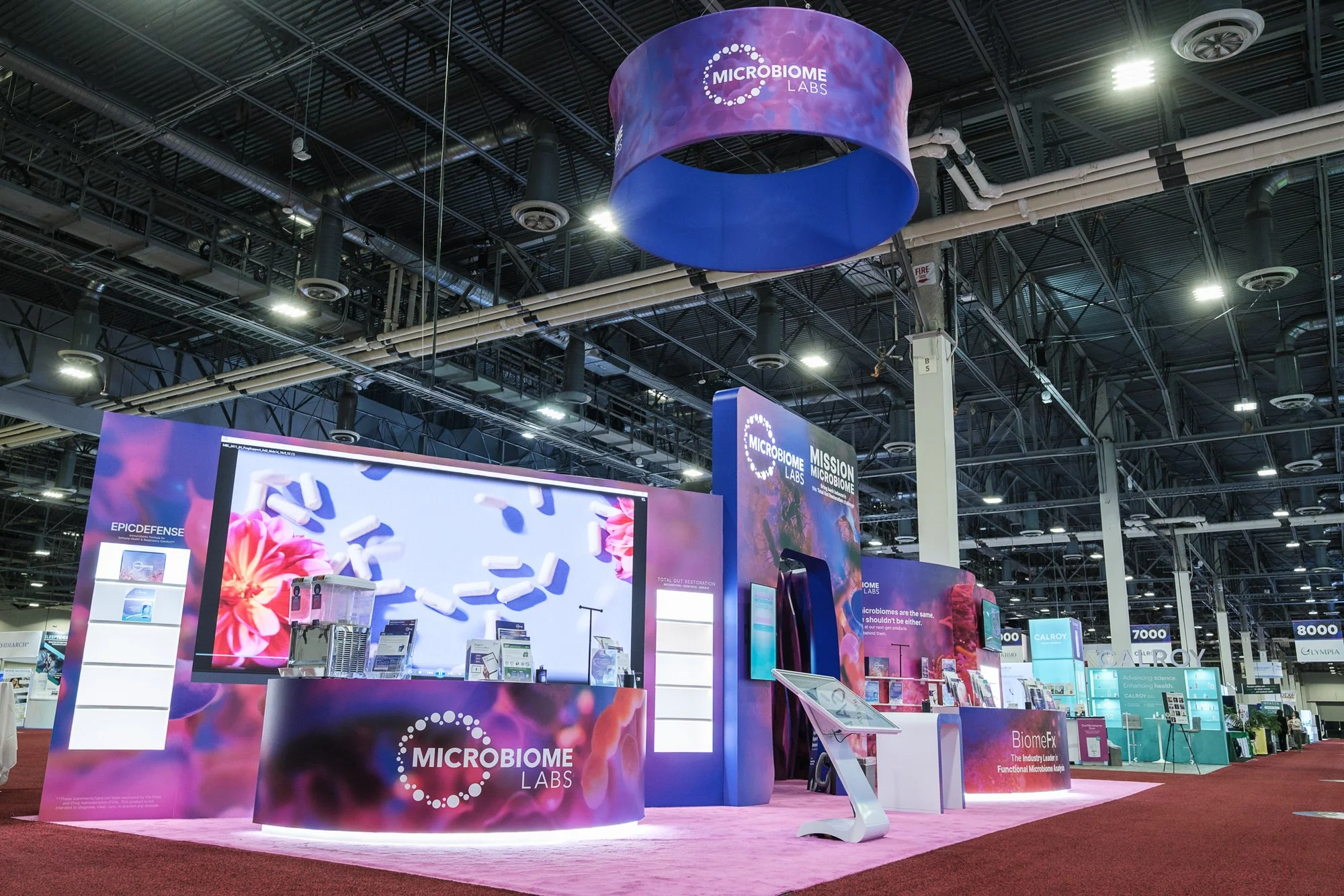 Exhibition booth for Microbiome Labs at a trade show. The booth features colorful graphics with molecular imagery and the company's logo. Overhead signage displays the company's name, and husky screens showcase information.