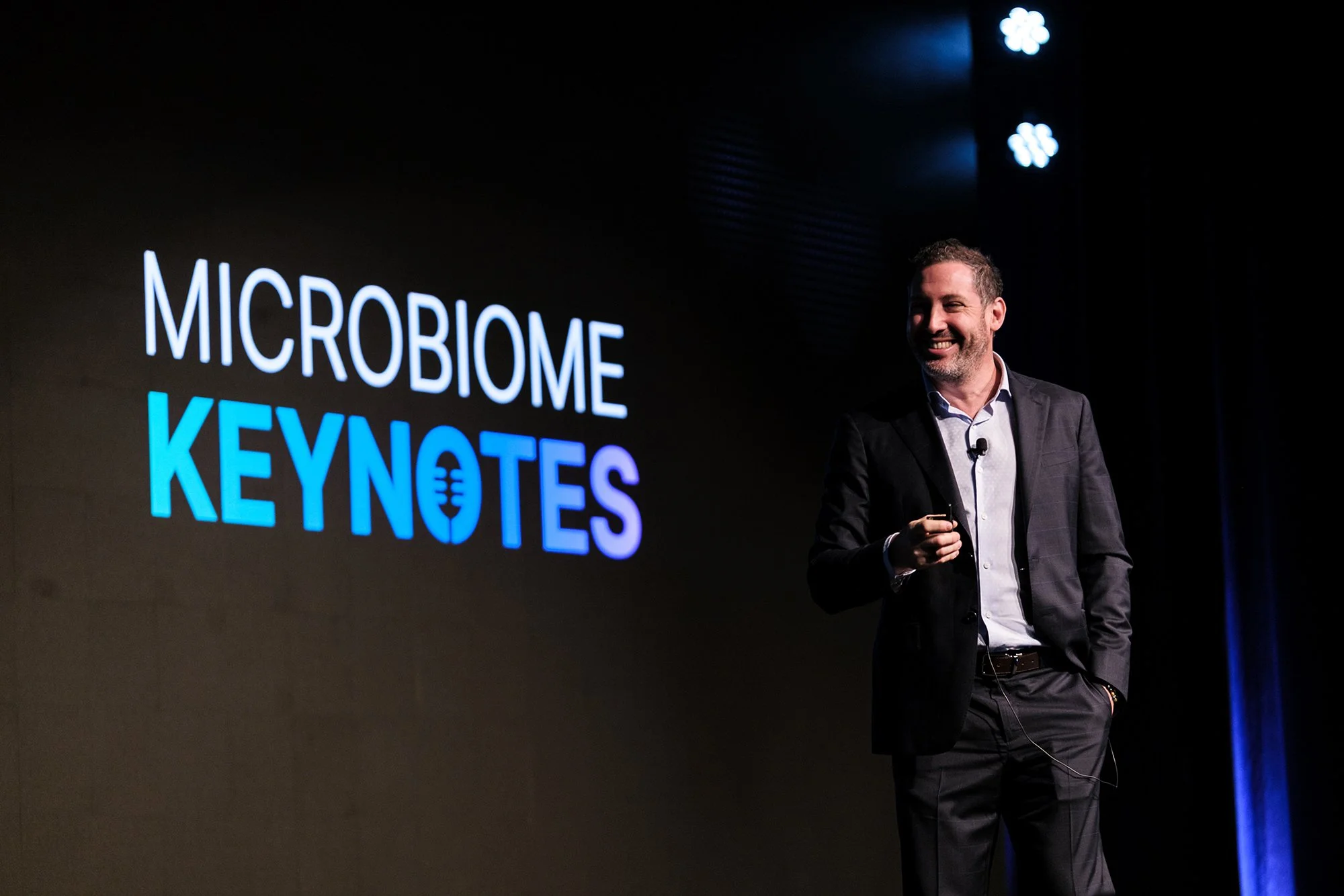 A man smiling and standing on stage in front of a large screen that reads 'MICROBIOME KEYNOTES' at a conference or presentation.