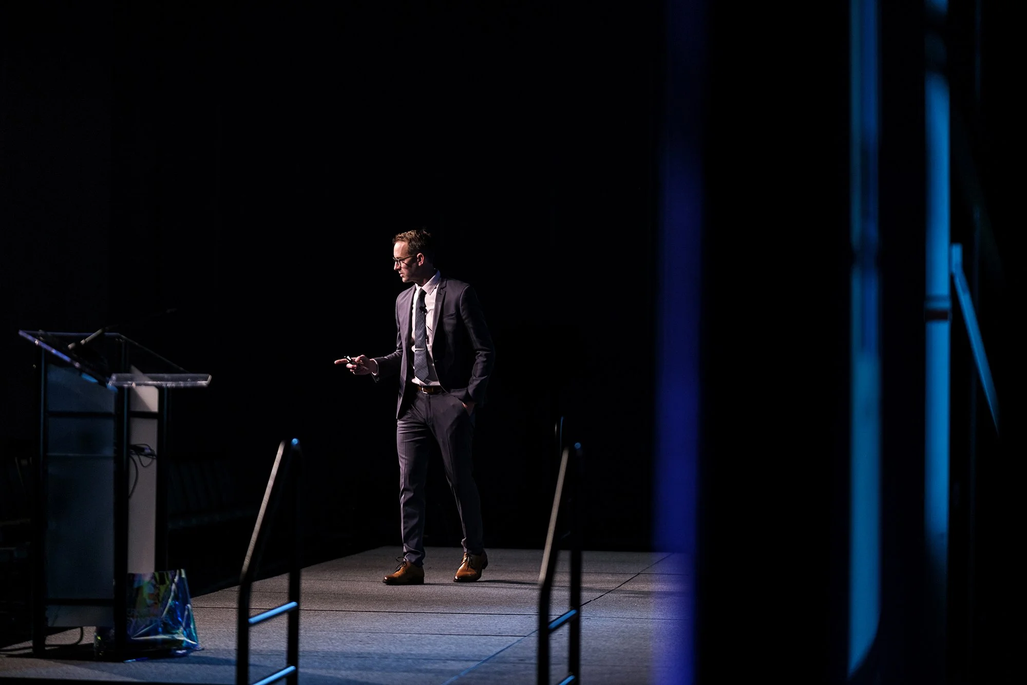 A man in a suit and glasses standing on a stage, looking at his phone, with a podium and presentation slides nearby, in a dark conference room.