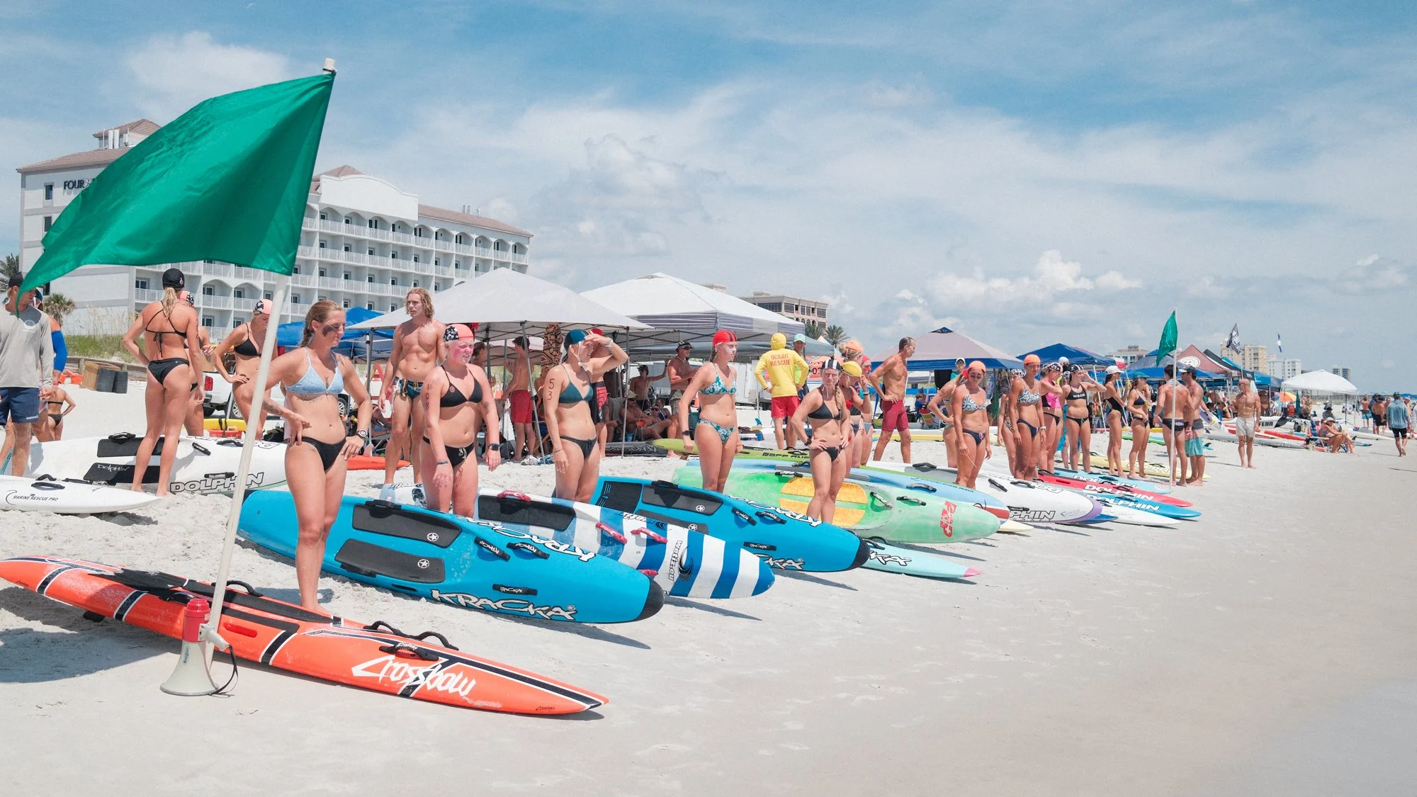 People at a beach with paddleboards and tents, some wearing swimsuits and swim caps, under a partly cloudy sky with buildings in the background.