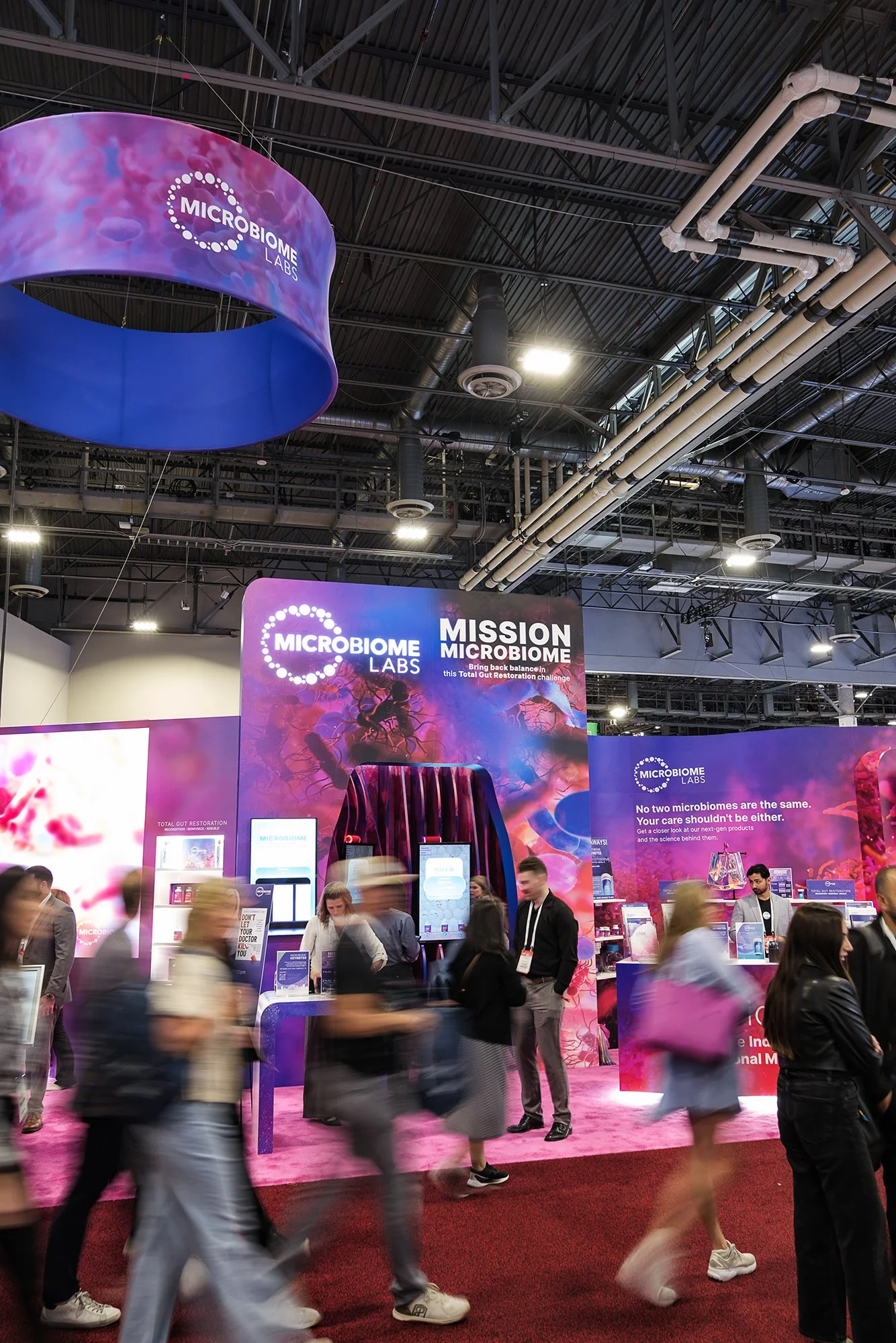Trade show booth for Microbiome Labs with signage about gut restoration, people walking around and engaging with displays, pink carpet flooring, industrial ceiling with pipes and lighting.