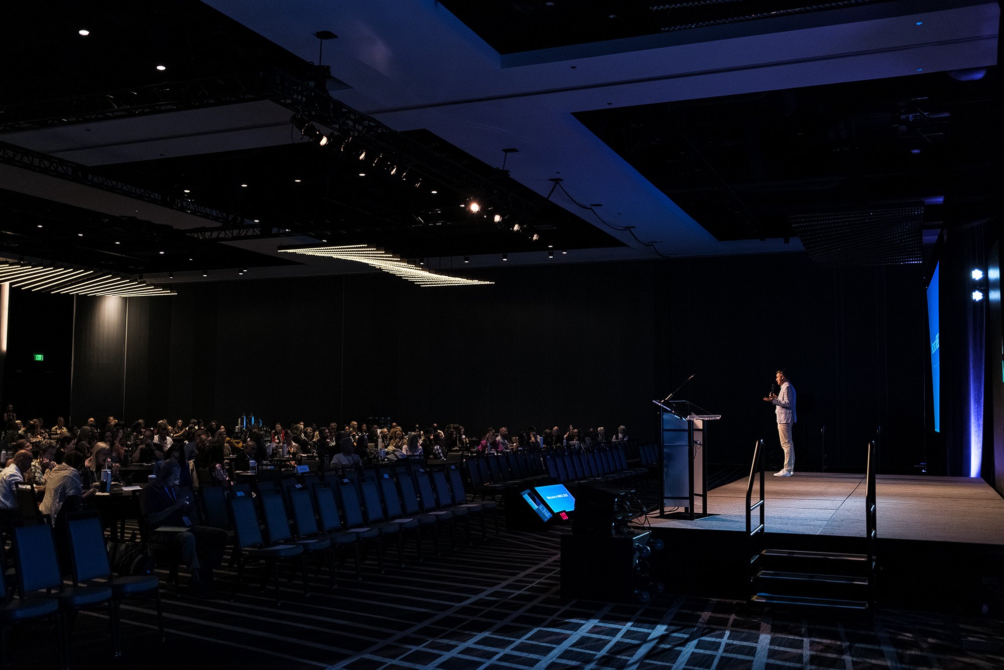 A conference room filled with rows of seated attendees watching a presenter on stage. The stage has a speaker at a podium, and stage lighting is directed towards the presenter. The ceiling features dark panels with multiple lights, and there are scre