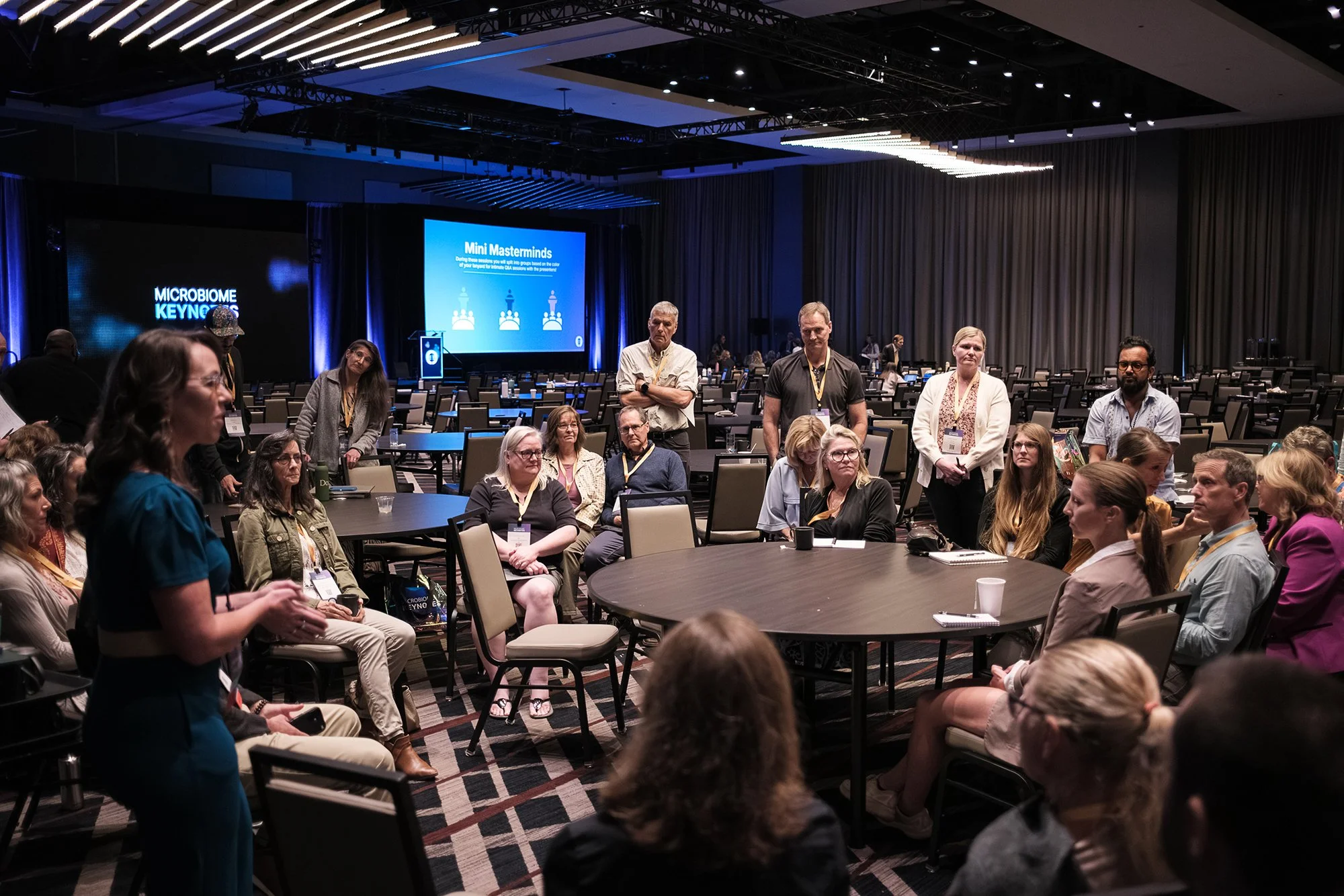 Conference attendees seated and standing in a large ballroom during a session, with a presentation slide titled 'Mini Masterminds' displayed on a screen in the background.