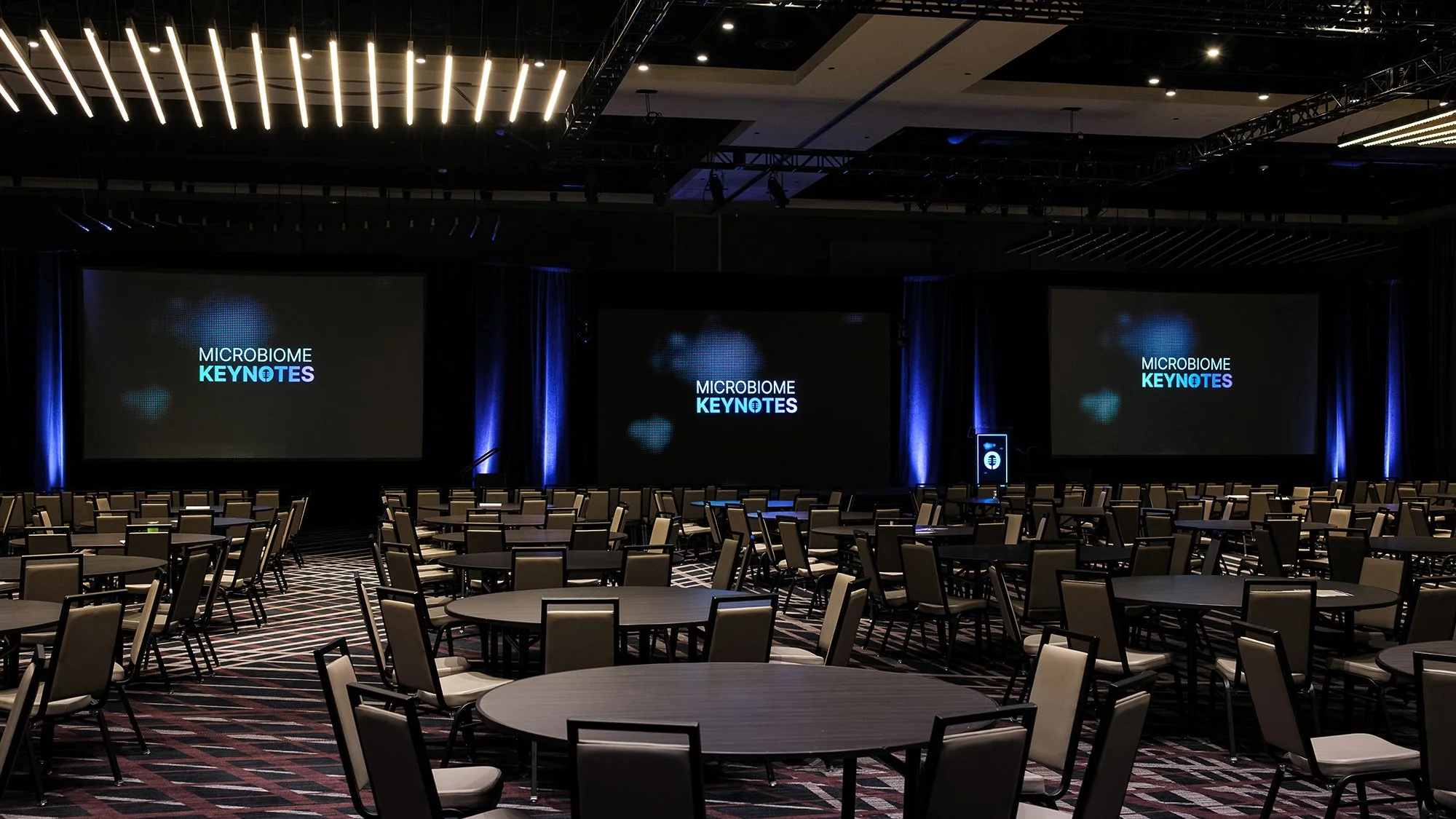 Conference room set up with round tables and chairs, large screens displaying 'MICROBIOME KEYNOTES,' and stage with podium, prepared for a presentation or event.
