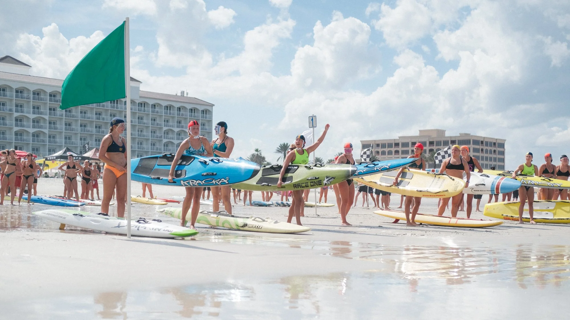 Women preparing for a paddleboarding race on the beach, holding paddleboards and wearing swimwear and caps, with a flagpole and buildings in the background under a partly cloudy sky.
