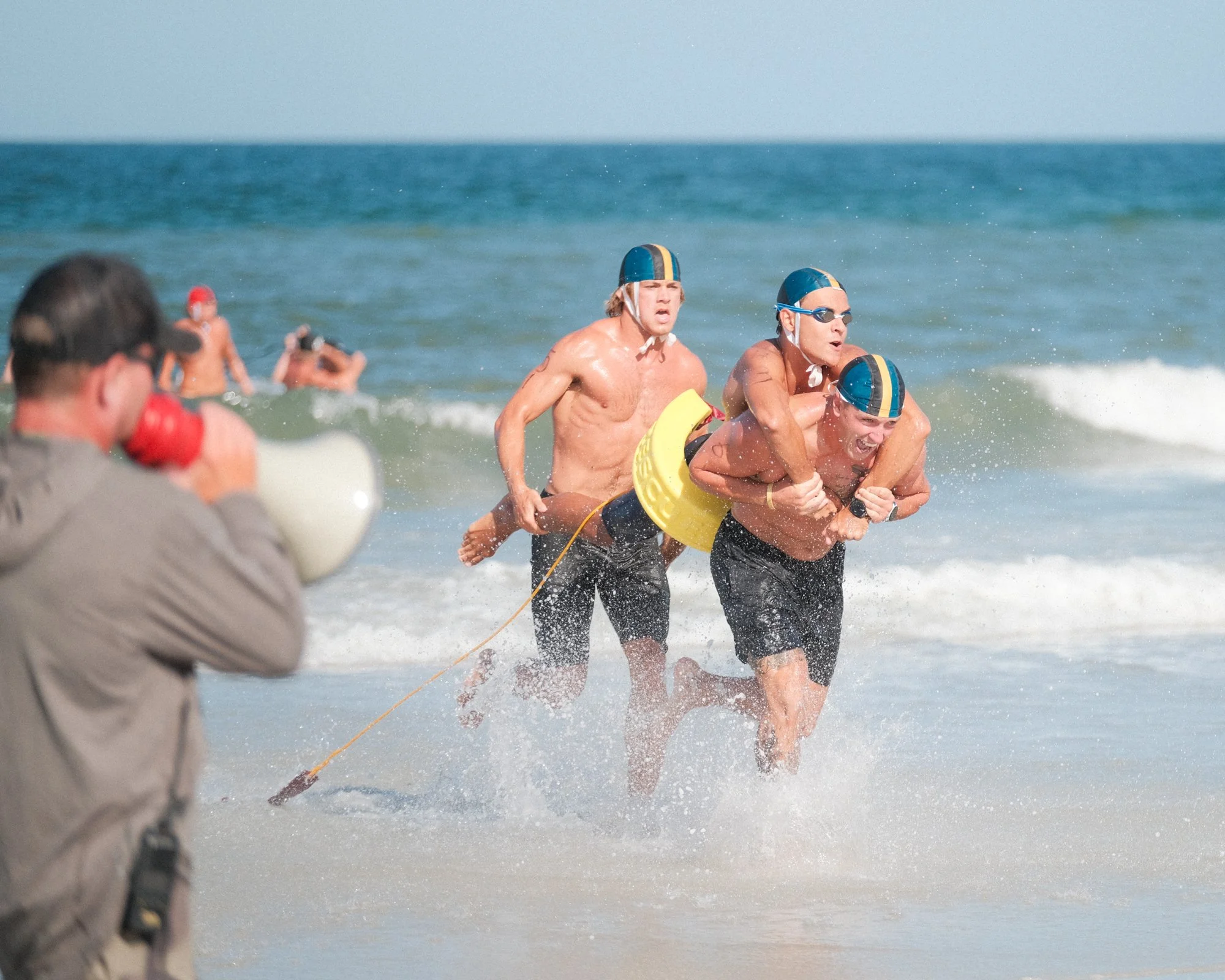 A group of men in swim caps participating in a rescue training or competition on the beach, with a photographer capturing the scene.