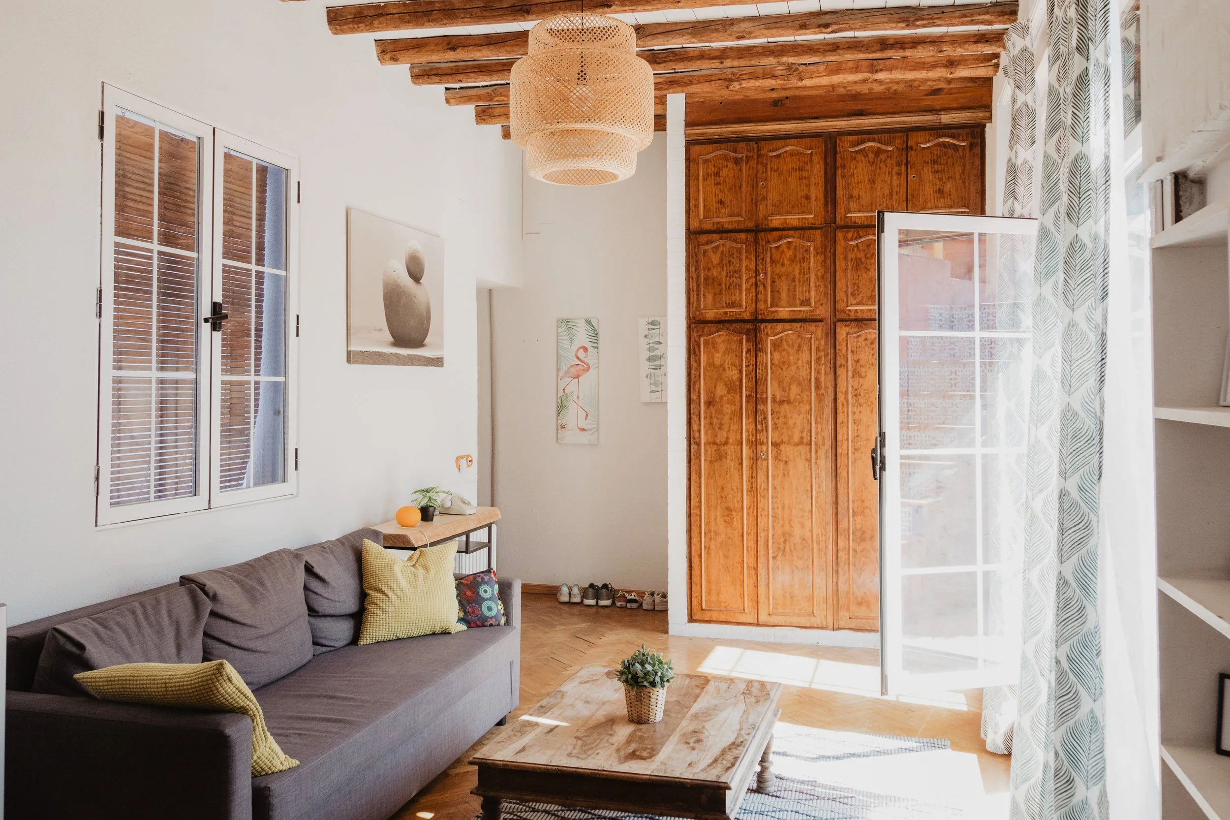 Professionally cleaned Living room with a dark gray sofa, patterned cushions, a wooden coffee table, a light fixture hanging from wooden beams, and a window with patterned curtains.