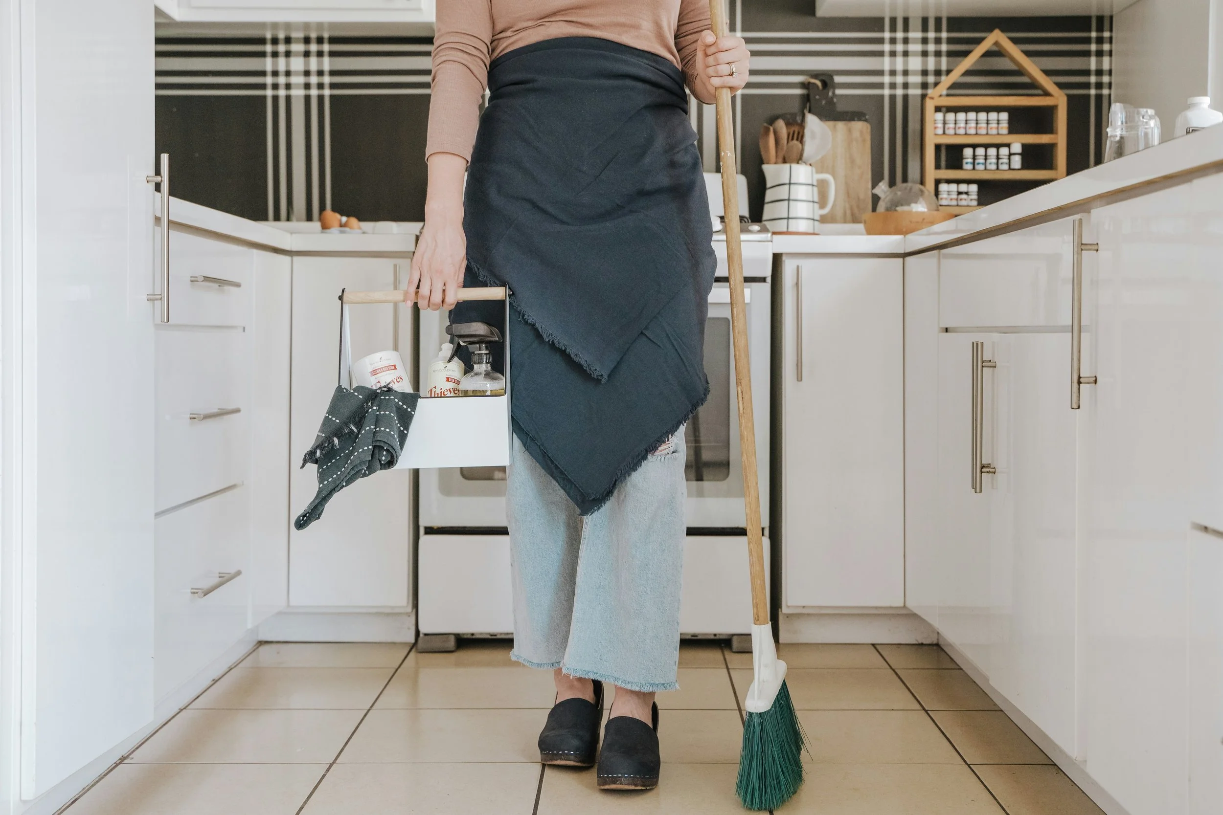 Person in a peach long sleeve shirt and jeans with a black apron holding a broom and a small cleaning caddy in a kitchen with white cabinets and tiled floor.