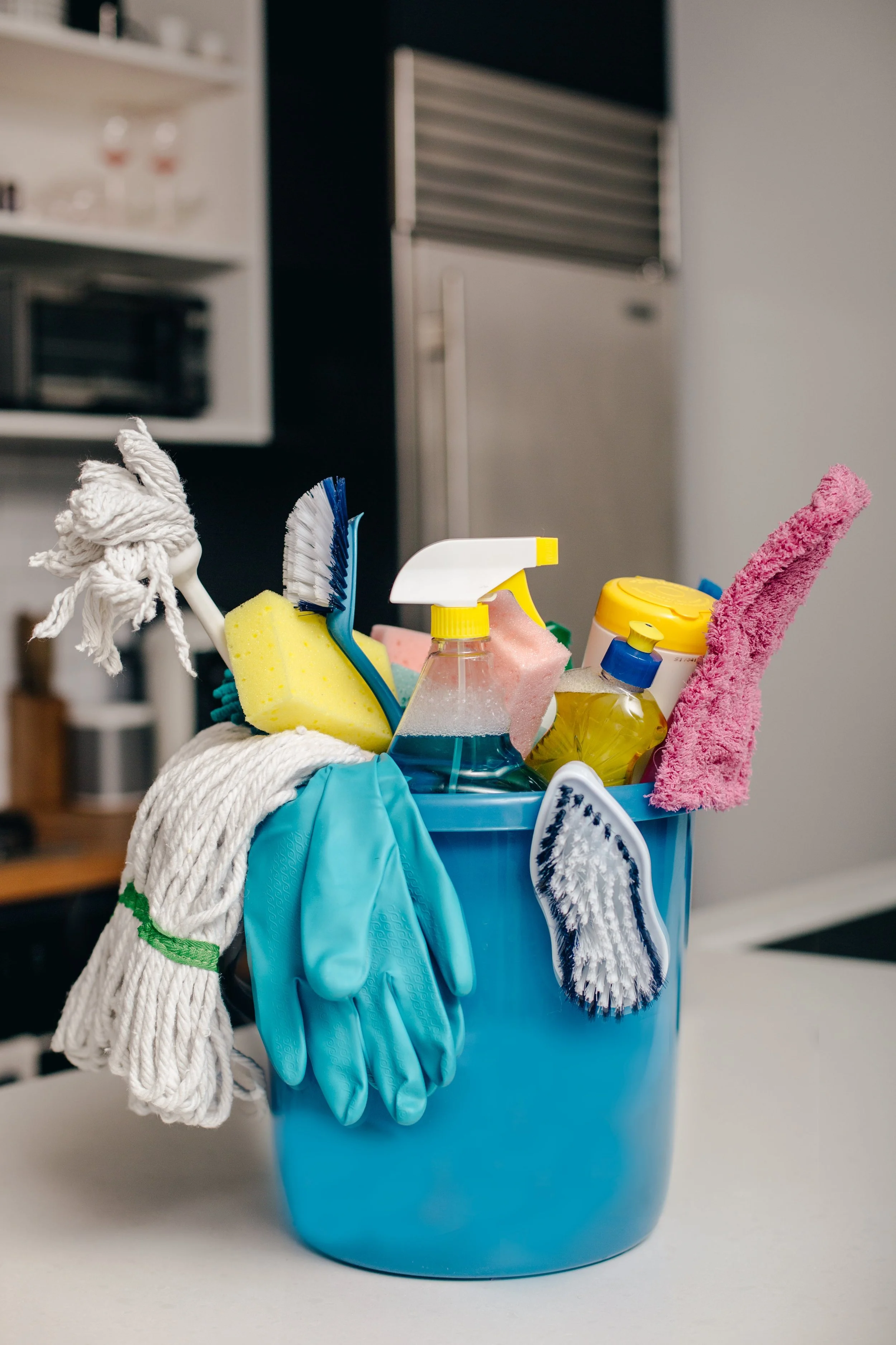A blue container filled with various cleaning supplies and tools, including sponges, spray bottles, gloves, and cleaning cloths, with a kitchen in the background. Housekeeper brings supplies