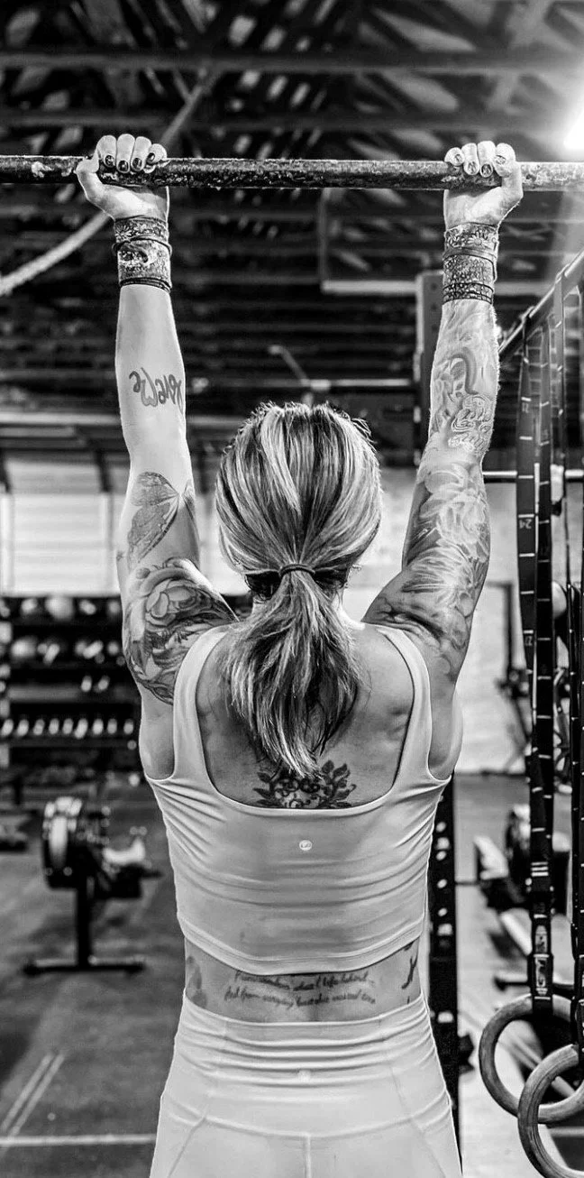 Tattooed woman performing a pull-up in a gym, seen from behind in black and white