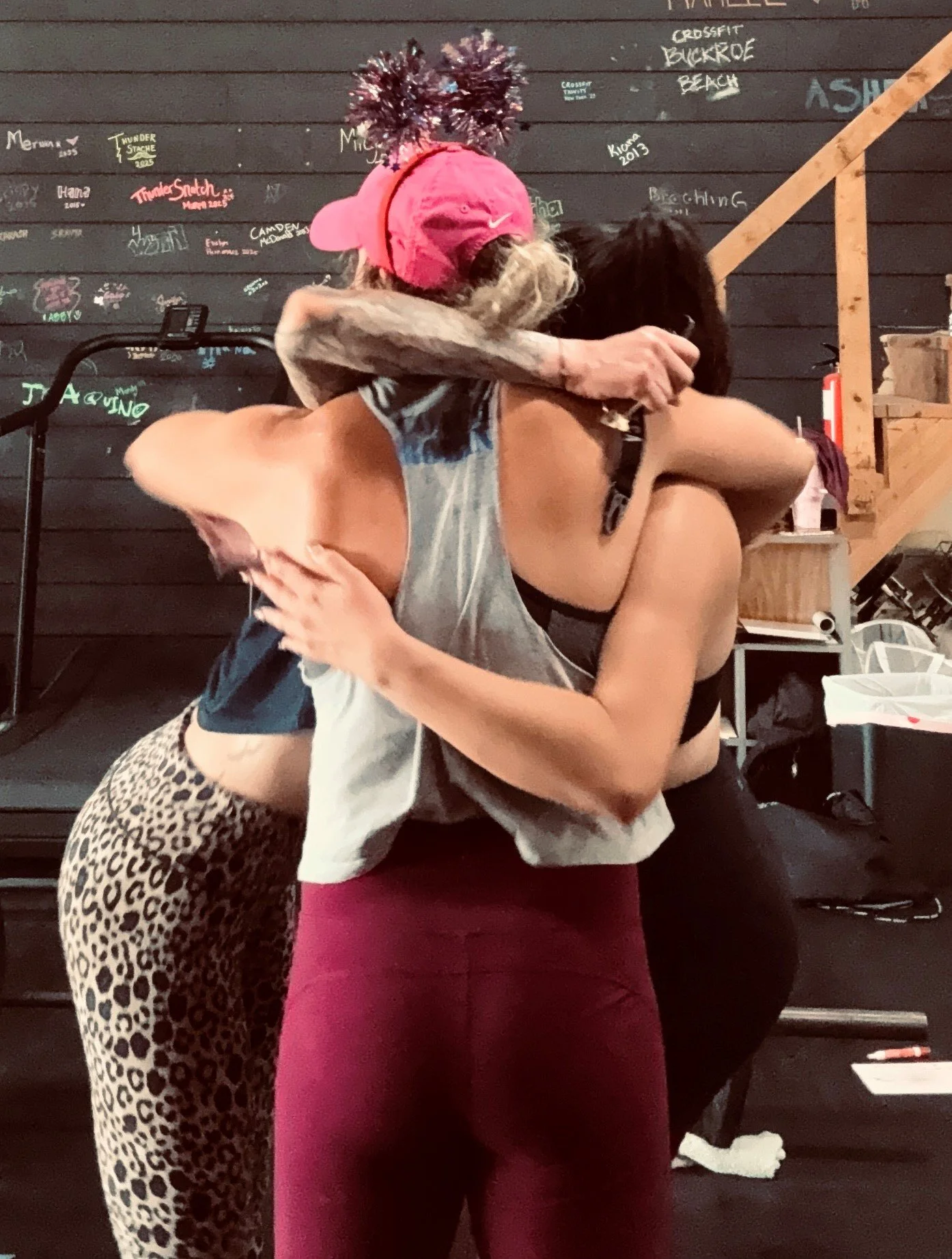 Two people hugging in a CrossFit gym, with a chalkboard workout wall and boxing equipment in the background.
