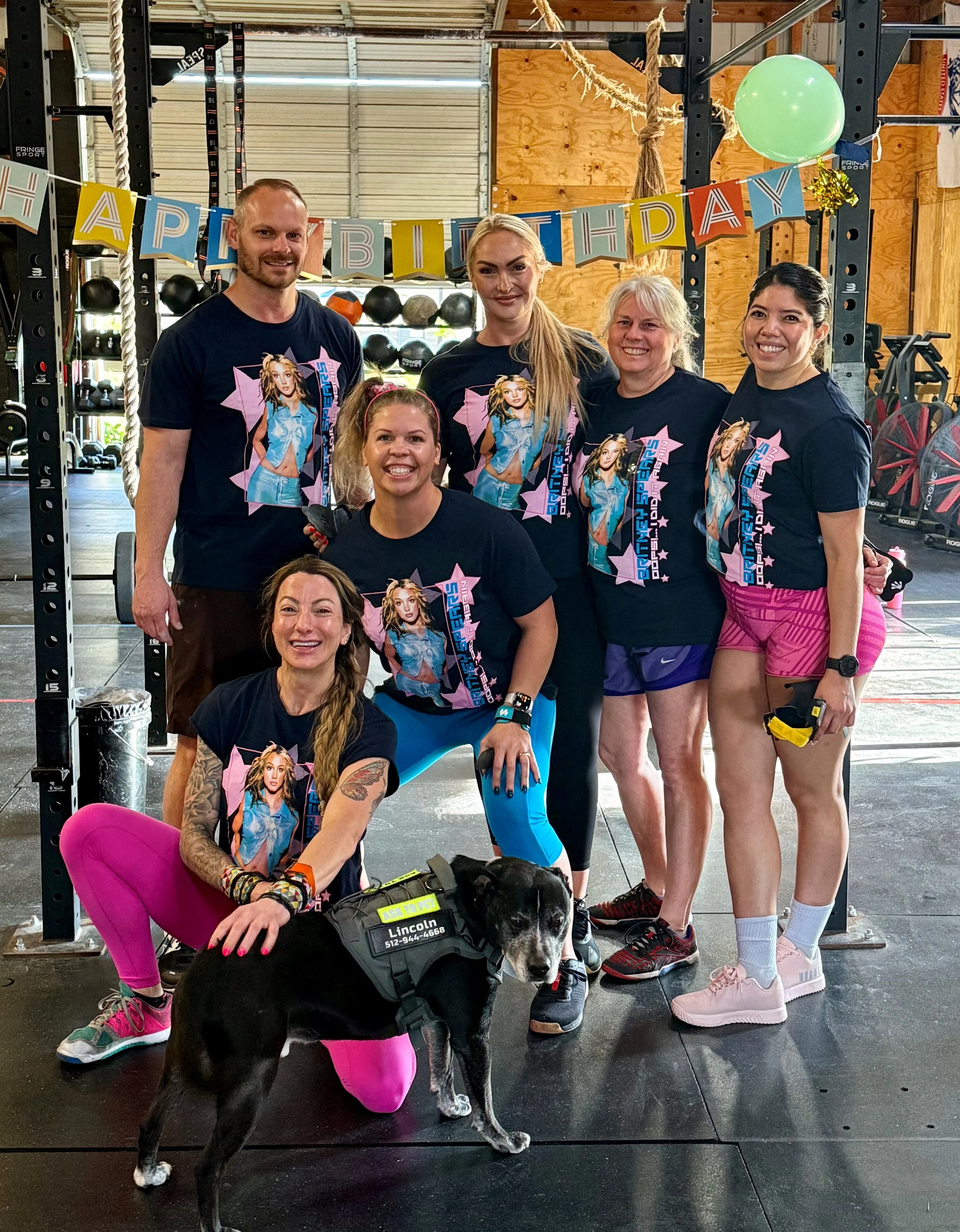 Group of people posing with a service dog in a decorated gym, wearing matching Britney Spears-themed shirts.