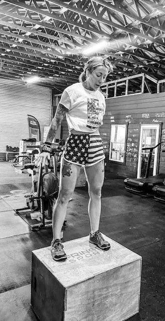 Black-and-white photo of a female athlete standing on a wooden plyometric box in a CrossFit-style gym.