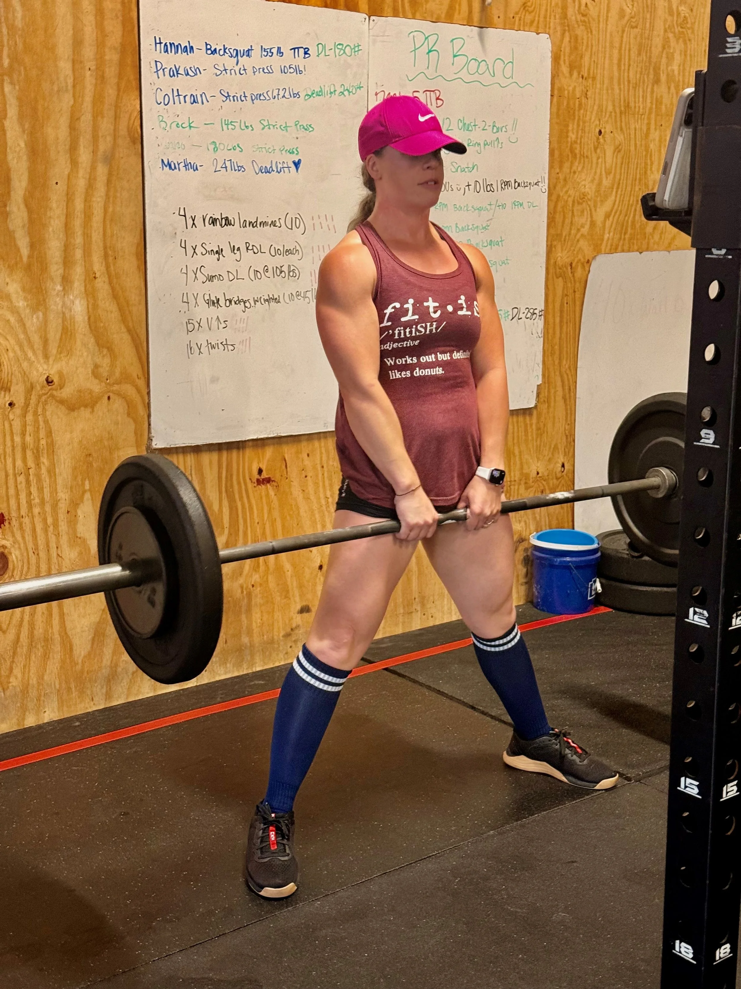 Woman in a pink cap and maroon tank top performing a barbell deadlift in a gym.