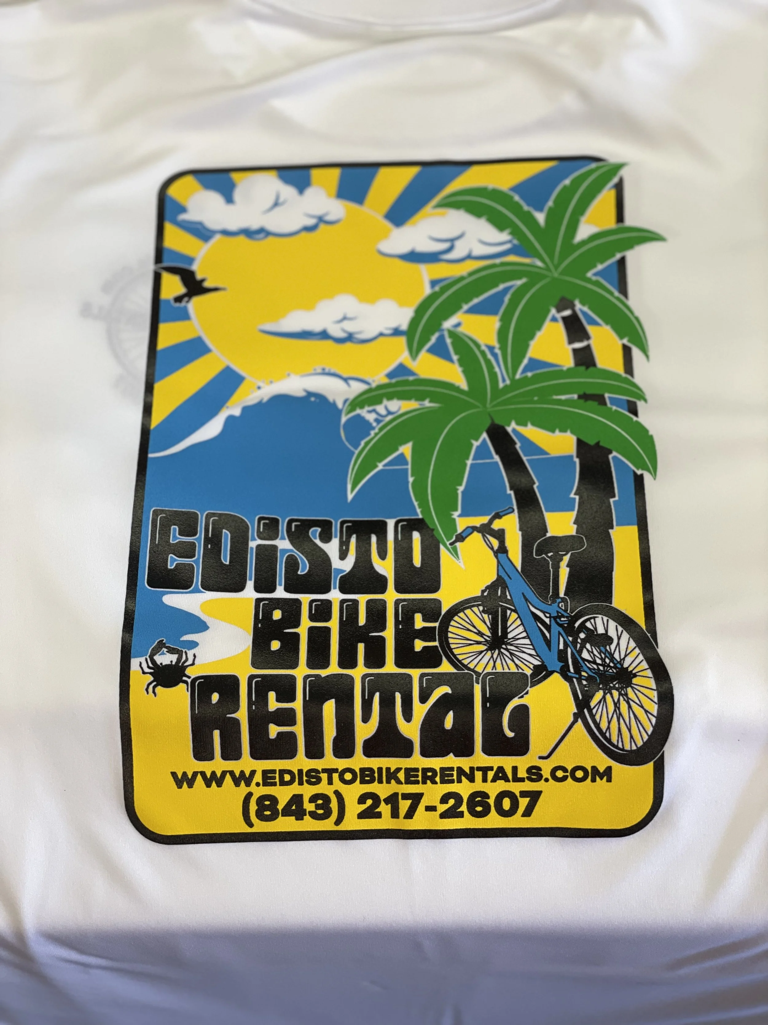 A colorful t-shirt design for Edisto Biker Rentals featuring a sun with rays, palm trees, clouds, a bird, a crab, a bicycle, and the website and phone number for the rental service.