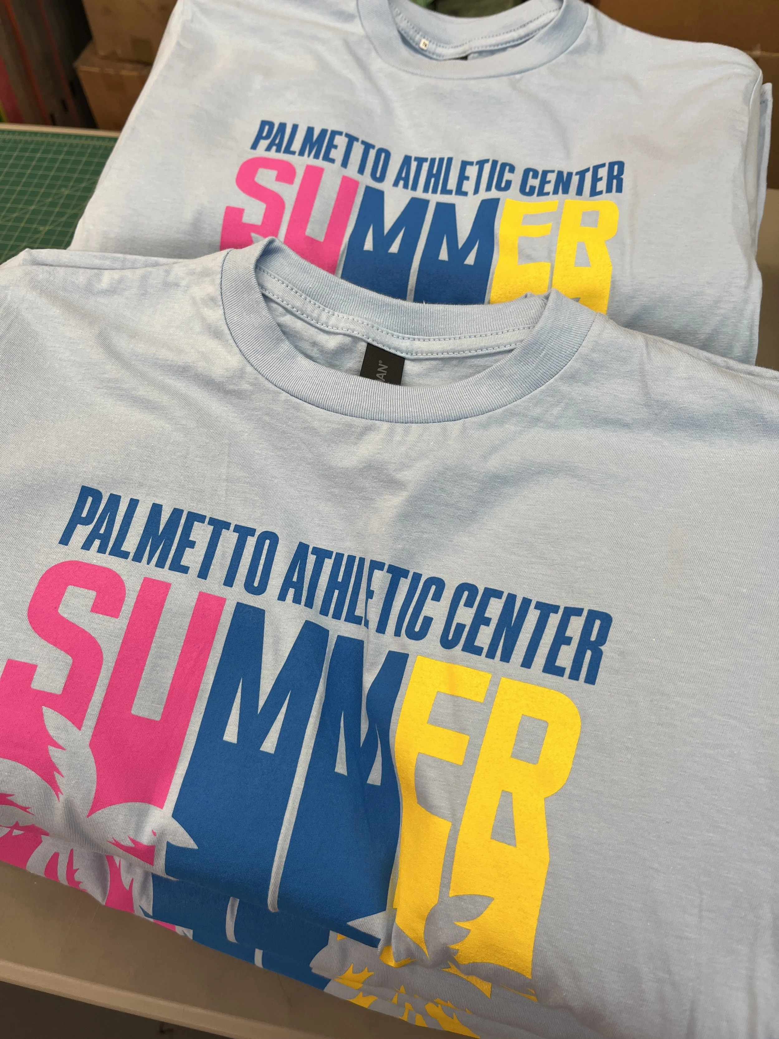 A stack of light blue T-shirts with colorful text for Palmetto Athletic Center Summer. The shirts display the words 'Palmetto Athletic Center' in blue, 'Summer' in pink, and 'Summer' in yellow with various designs and colors.