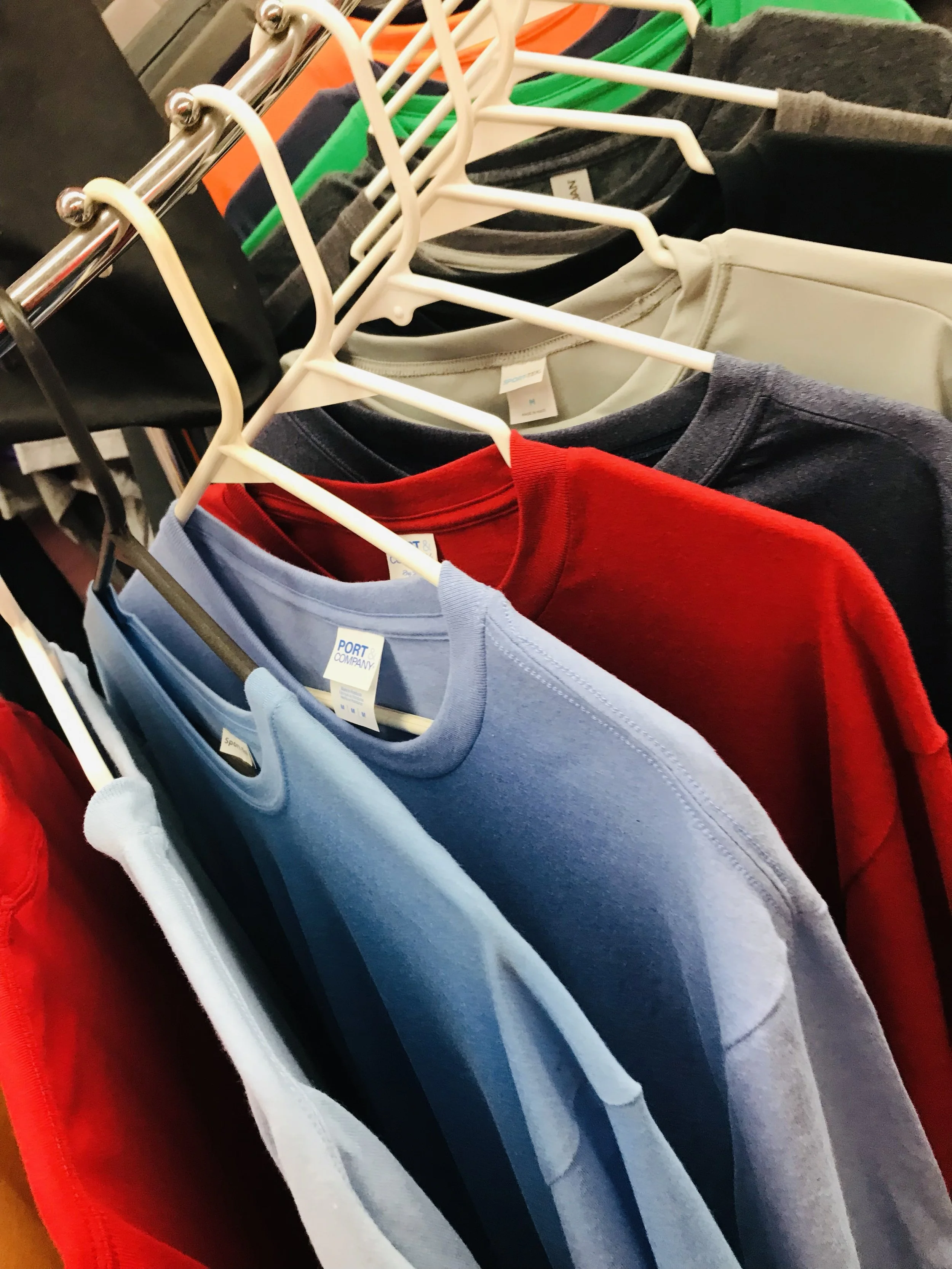 A clothing rack with colorful shirts on hangers, including light blue, red, navy, gray, black, green, orange, and beige, displayed in a t-shirt shop.