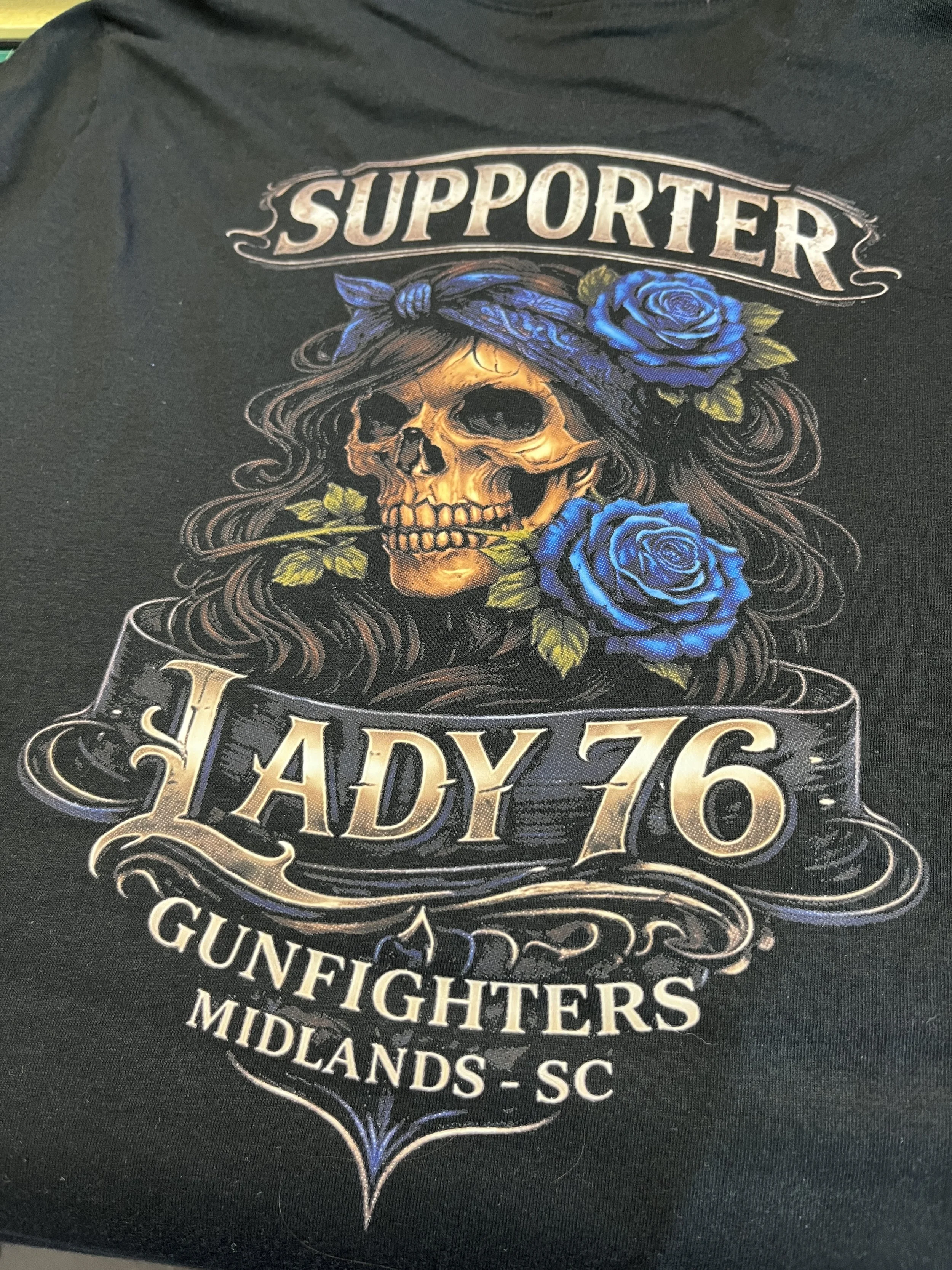 A Direct To Film (DTF) transfer on dark t-shirt showing a skull with long hair, a bandana, and blue roses. The graphic includes the words 'SUPPORTED LADY 76' and references motorcycle clubs and locations in South Carolina.