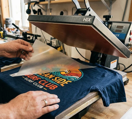 A person using a heat press machine to apply a colorful Direct To Film Transfer DTF logo design onto a navy blue T-shirt.
