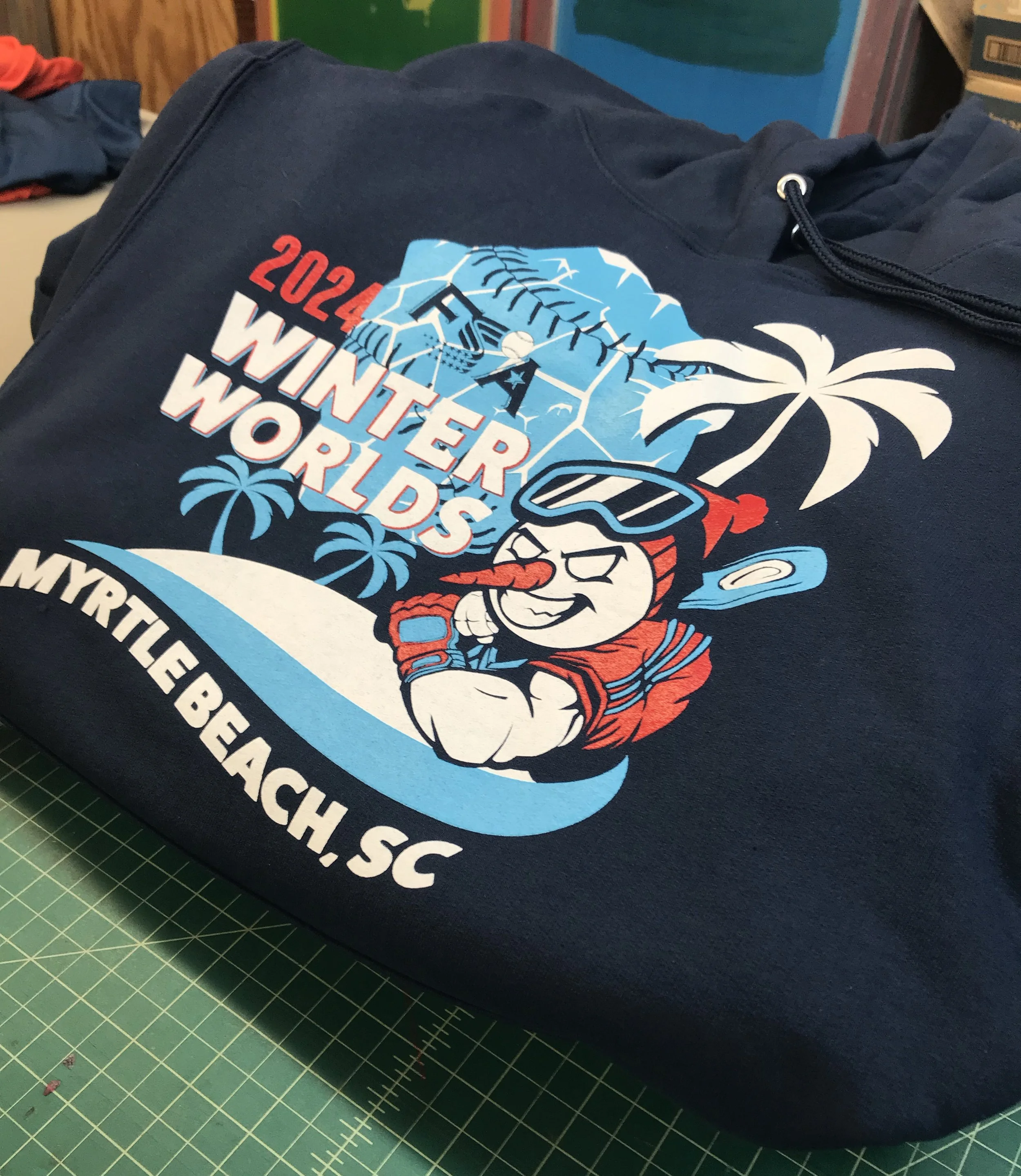 Close-up of a custom screen printed navy blue T-shirt featuring a design with the text 'FSA 2024 WINTER WORLD SERIES' and an illustration of a smiley face with sunglasses and a baseball cap, surrounded by palm trees and a mountain.