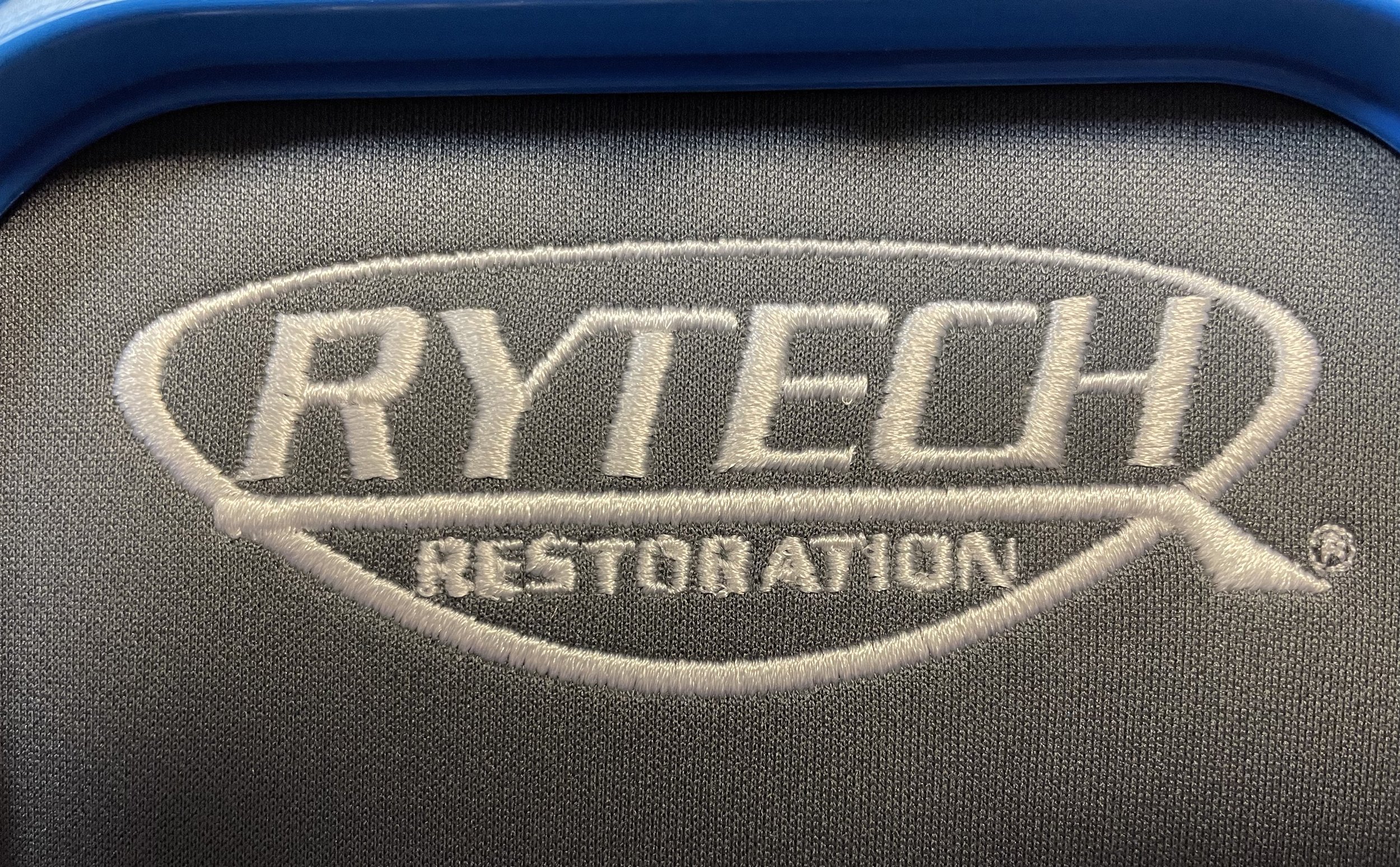 Close-up of embroidery on fabric, displaying the words "RYTECH RESTORATION" inside a shield-shaped logo with a gray background.