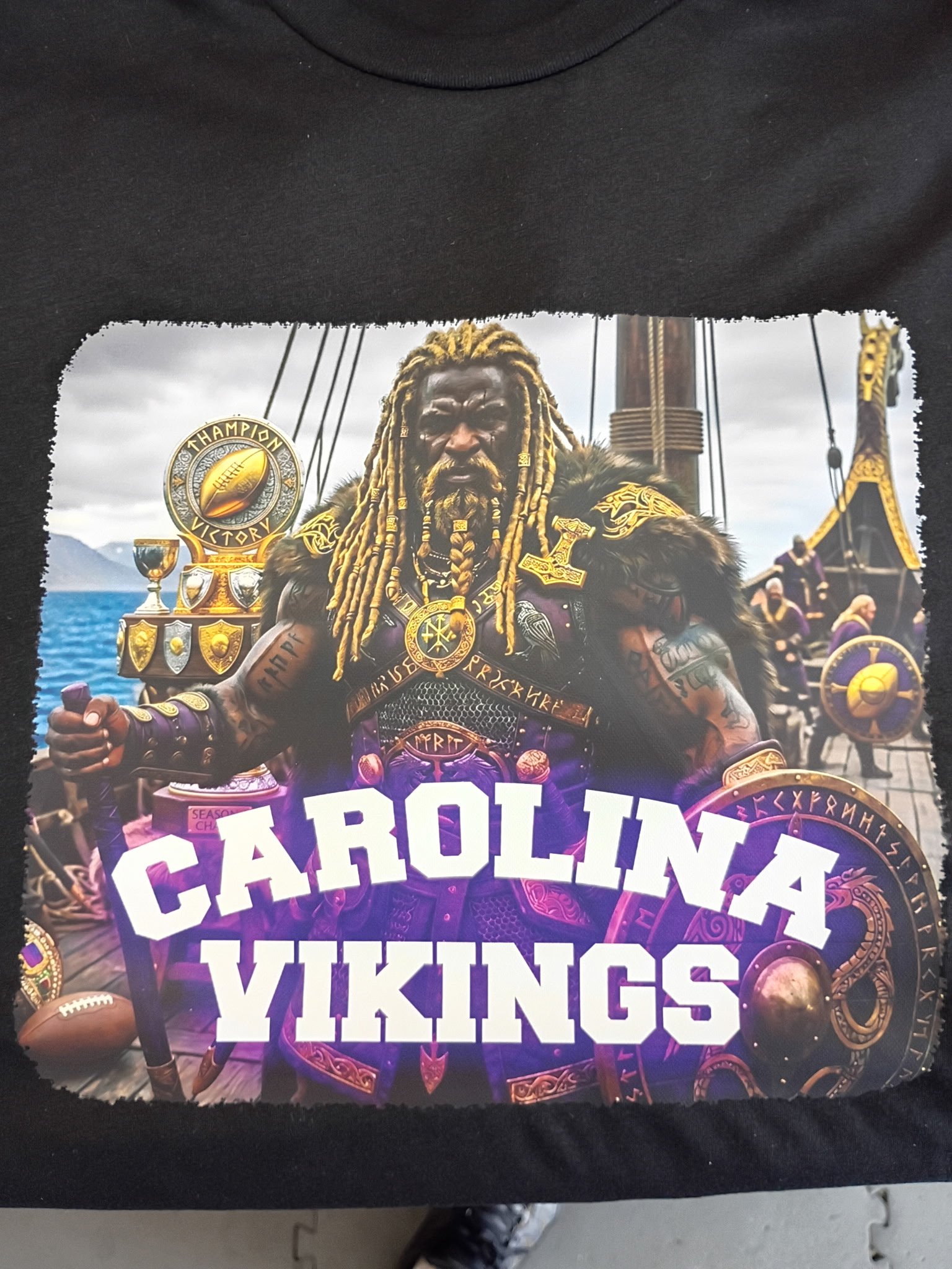 A graphic T-shirt featuring a Viking warrior with long blonde dreadlocks, wearing armor, and holding a shield. The background depicts Viking ships with sails. The shirt has bold white text saying 'CAROLINA VIKINGS'.
