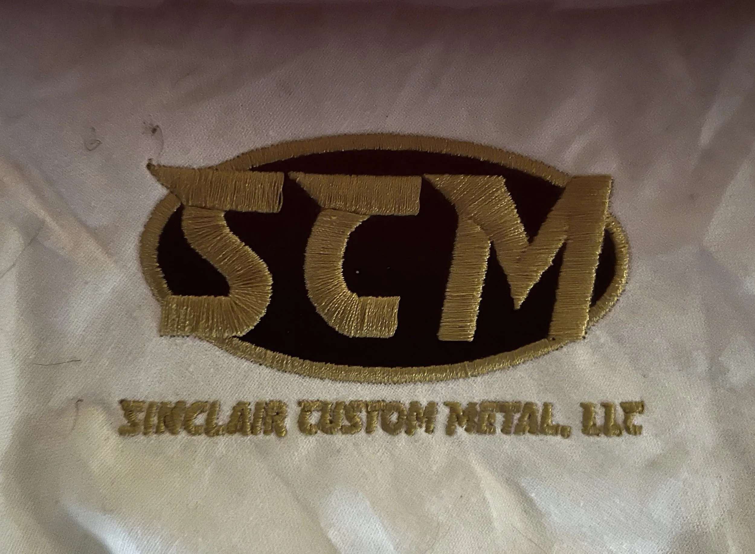 Close-up of embroidery on white fabric displaying the logo of SCM Sinclair Custom Metal LLC.