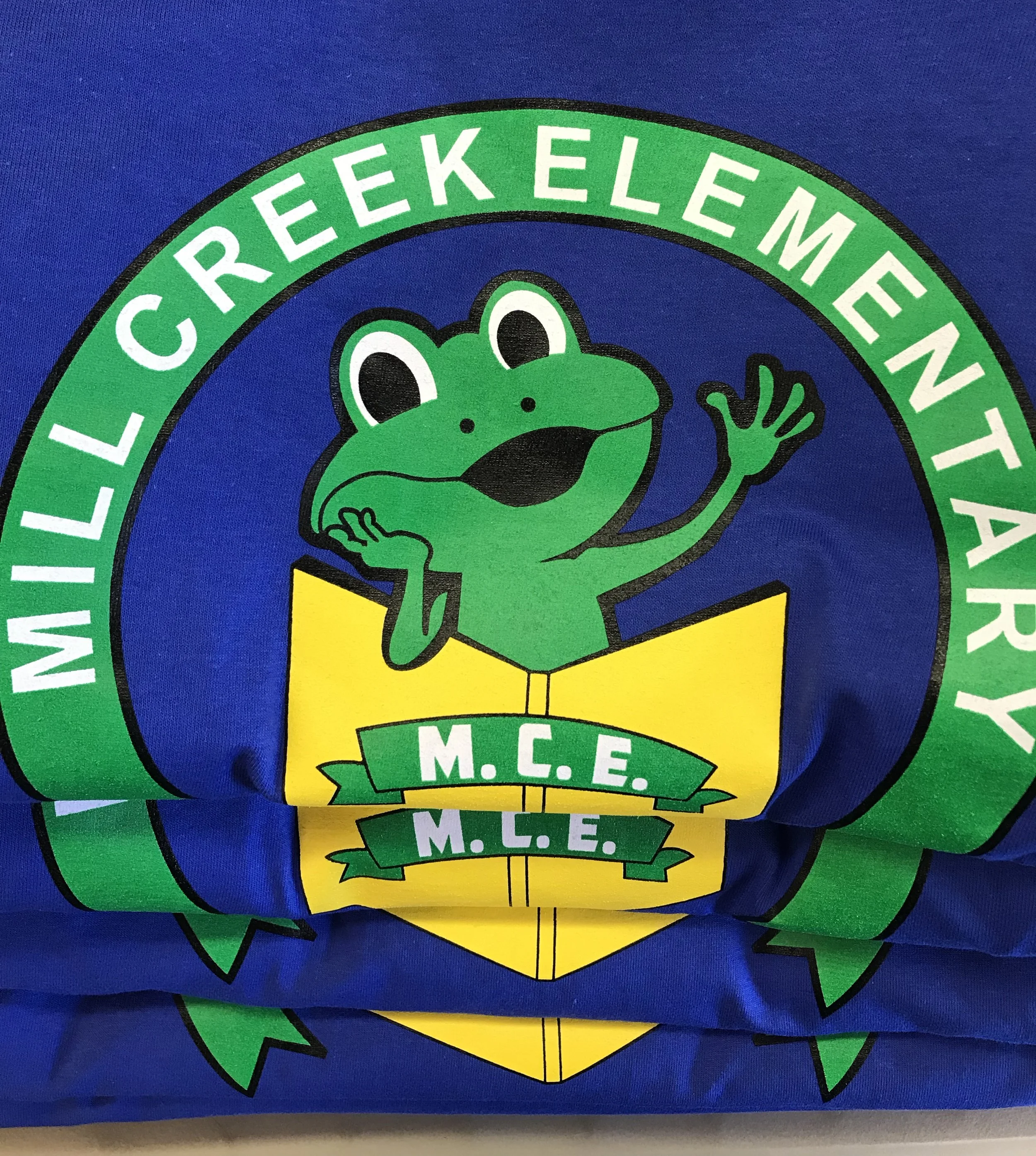 A screen printed design on a royal t-shirt, featuring a cartoon green frog wearing a yellow shirt. The frog is smiling and waving. The logo has a green and white outer ring with the text 'Mill Creek Elementary.' printed on it.