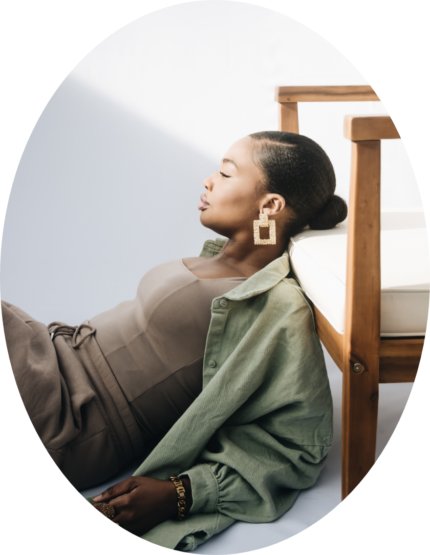 A woman with her eyes closed, reclining against a bed, wearing beige and green clothing and large square earrings.