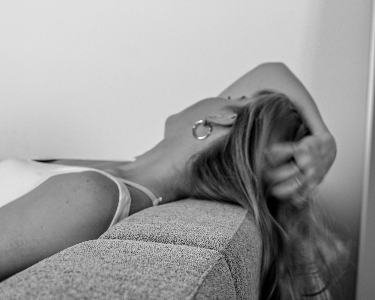 Black and white photo of a woman lying on a couch with her head resting on the armrest, her hand in her hair, wearing a tank top and hoop earrings.