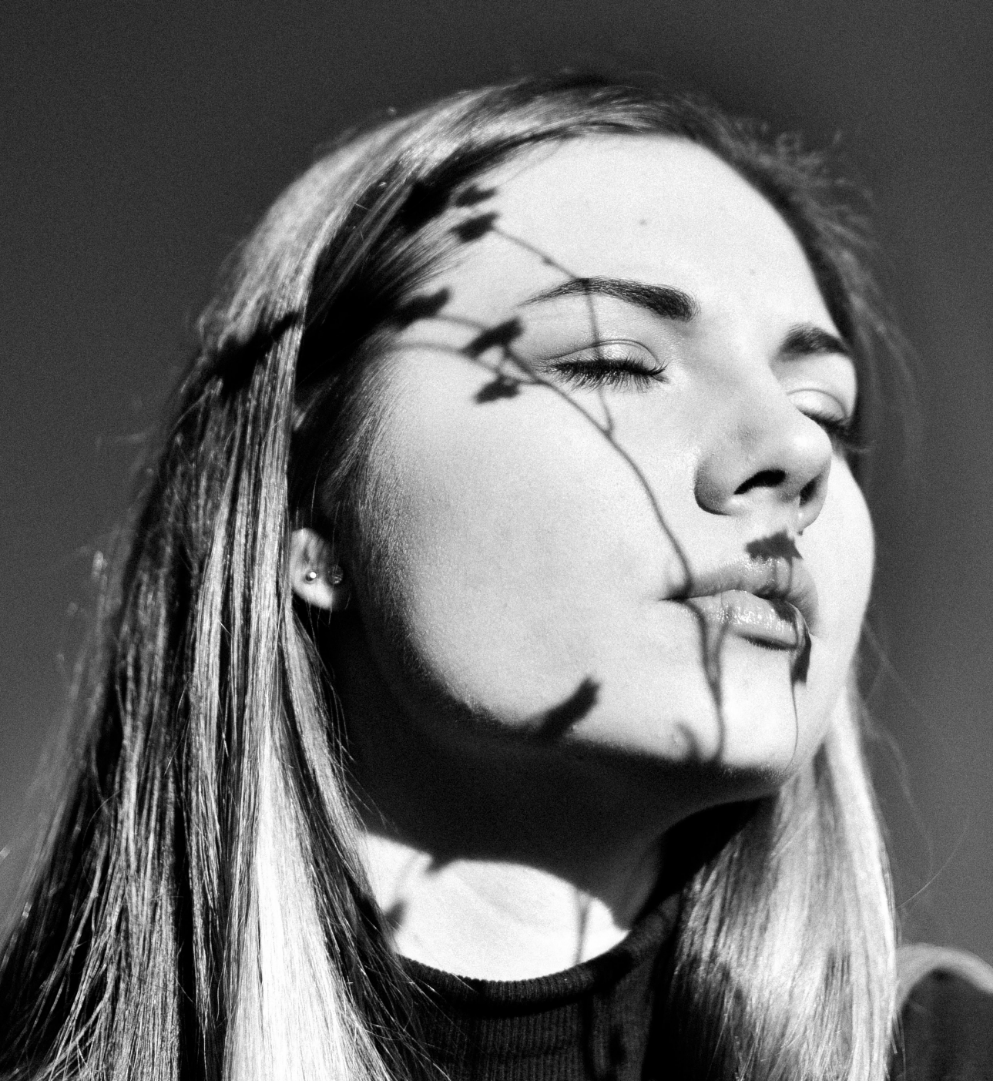 Black and white photo of a woman with long hair, eyes closed, with a shadow of a plant branch on her face.