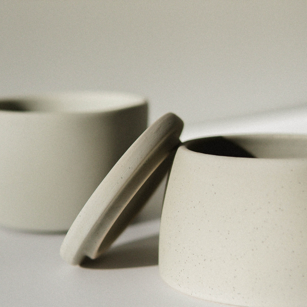 Close-up of two minimalist ceramic bowls, one with a lid, in neutral tones on a light background.