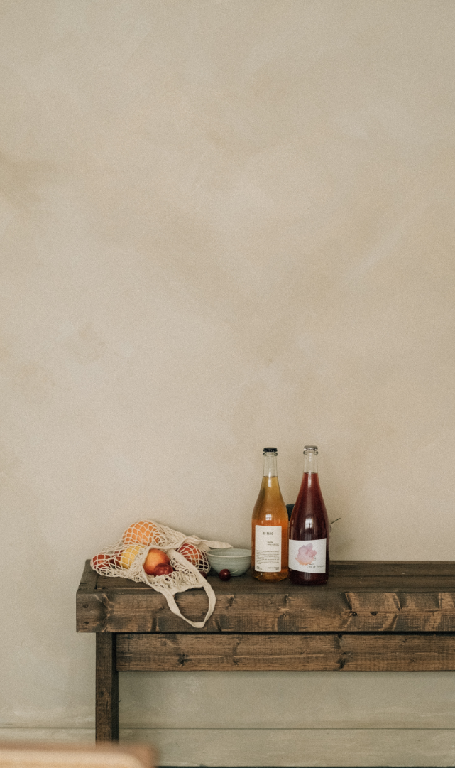A wooden table against a beige wall with two bottles, a bowl, a plastic bag with tomatoes and peaches, and a cherry.
