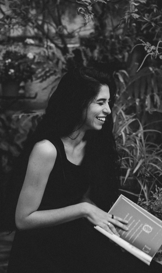 A woman with long dark hair smiling and holding a book, outdoors near plants.