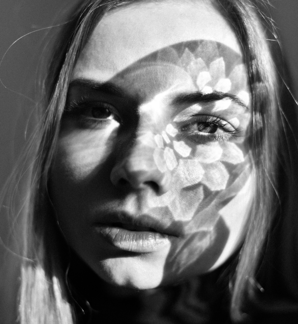 Close-up black-and-white photo of a woman’s face with shadows of leaves and flower patterns cast on her face.