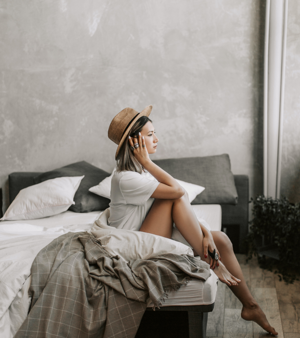 A woman sitting on the edge of a bed, wearing a white dress and a wide-brimmed hat, listening to music with headphones in a bedroom with gray walls and a gray headboard.