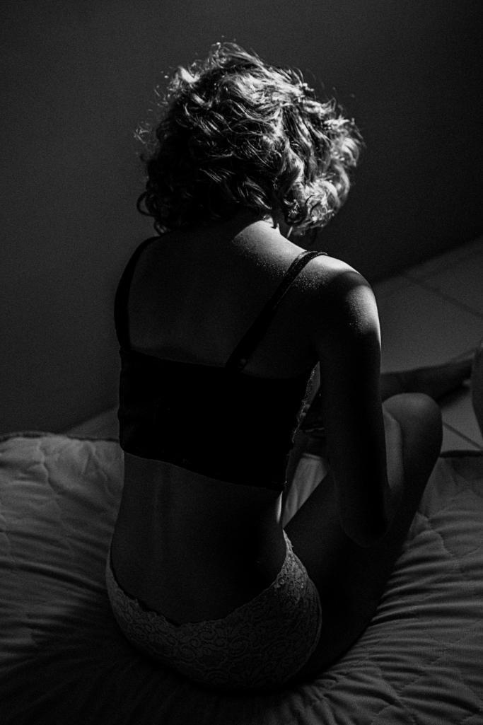 A person with curly hair sitting on a bed in black and white. The lighting highlights their shoulder and back, and they are wearing a tank top and lace underwear.